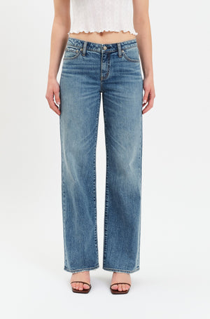 A person is shown from the torso down, wearing Daze's The Keeper Low Rise In Heartbeat Vintage jeans—a blue comfort stretch denim wide-leg style—paired with a white sleeveless crop top and black heeled sandals, standing against a plain white background.