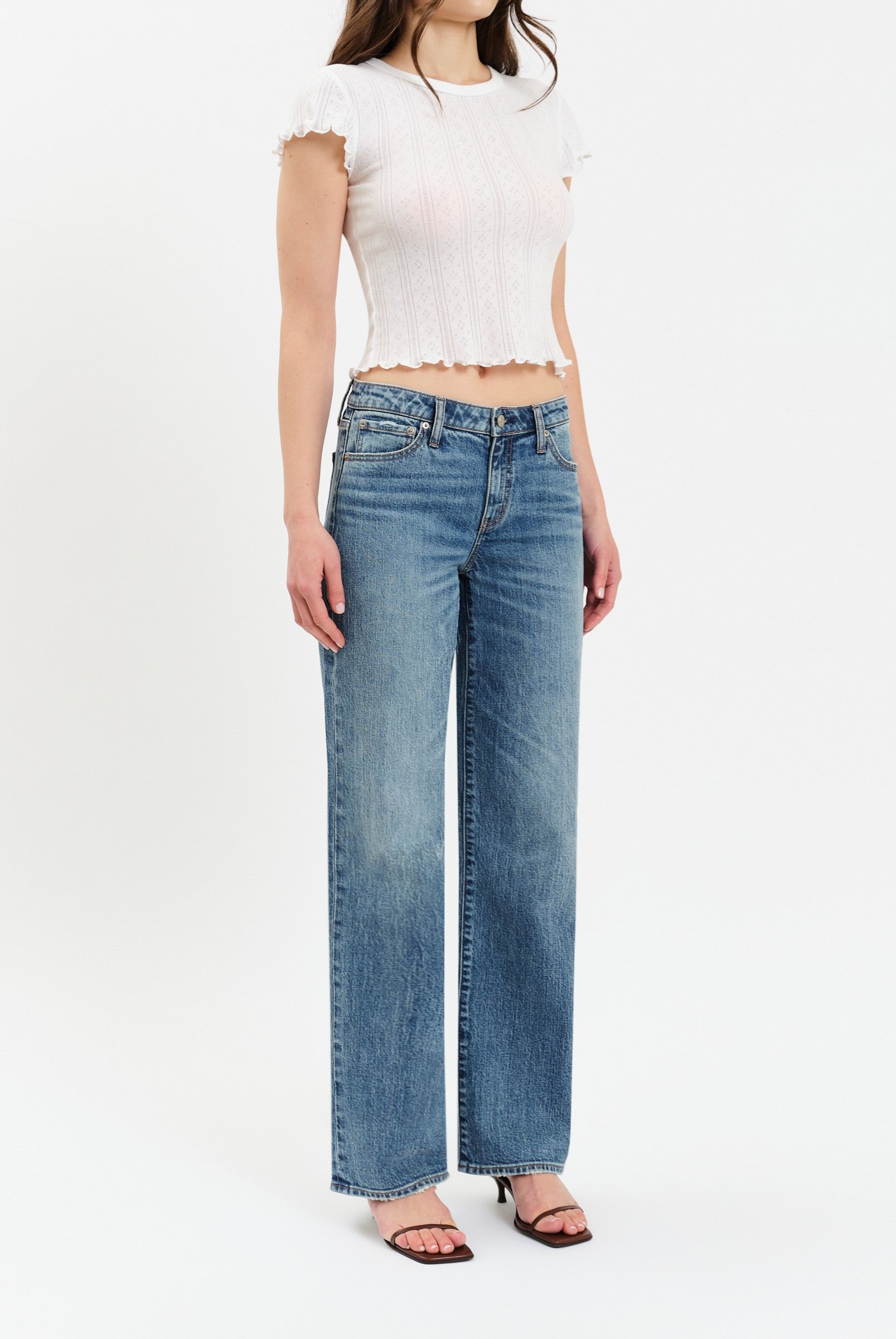 A person wearing a white short-sleeve crop top, The Keeper Low Rise In Heartbeat Vintage jeans by Daze, and black square-toe sandals stands against a plain white background.