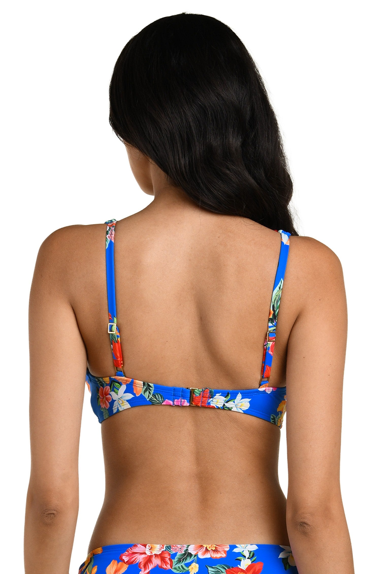 A woman with long dark hair, seen from the back, wears the La Blanca Modern Oasis Multi Ots Tri bikini top in bright blue with a colorful floral print and adjustable shoulder and back straps.