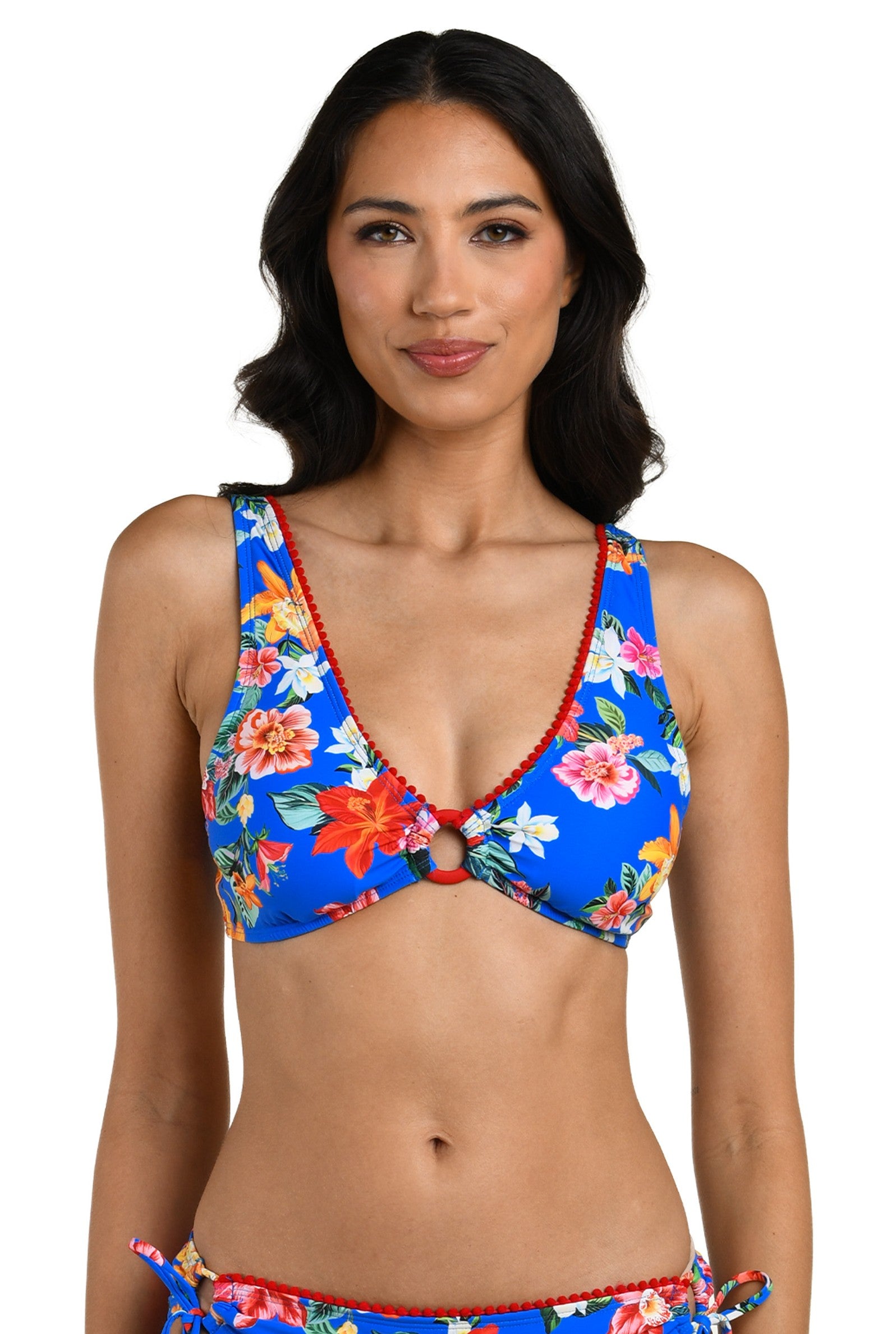A woman with medium-length dark hair models the La Blanca Modern Oasis Multi Ots Tri bikini top, featuring a blue floral print, red trim, small front ring detail, and adjustable straps. She smiles softly against a plain white background.