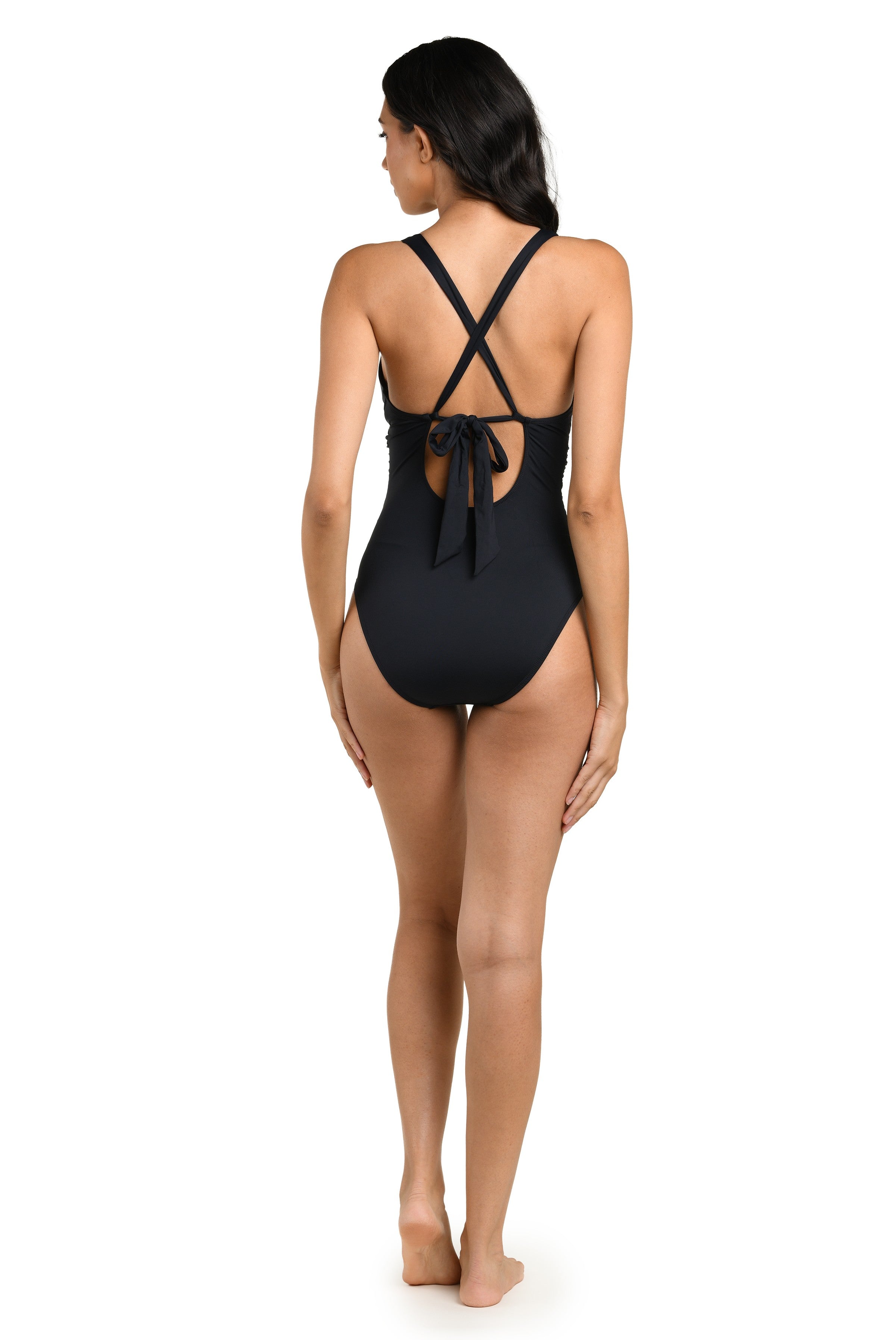 A woman, barefoot and facing away, models the La Blanca Black Soft Strap Twist Front Mio swimsuit with crisscross straps, moderate coverage, and a tied bow at the lower back. Her dark hair is loose; nylon elastane. Hand wash recommended.