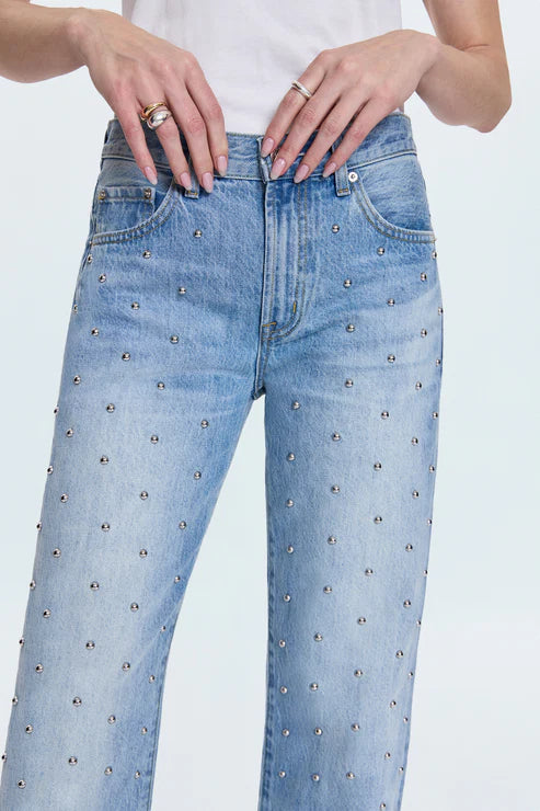 A person wearing Pistola's Lexi In Confetti high-waisted, light blue 100% cotton jeans with small silver studs and a relaxed fit, paired with a white top. Only the lower torso and hands are visible.