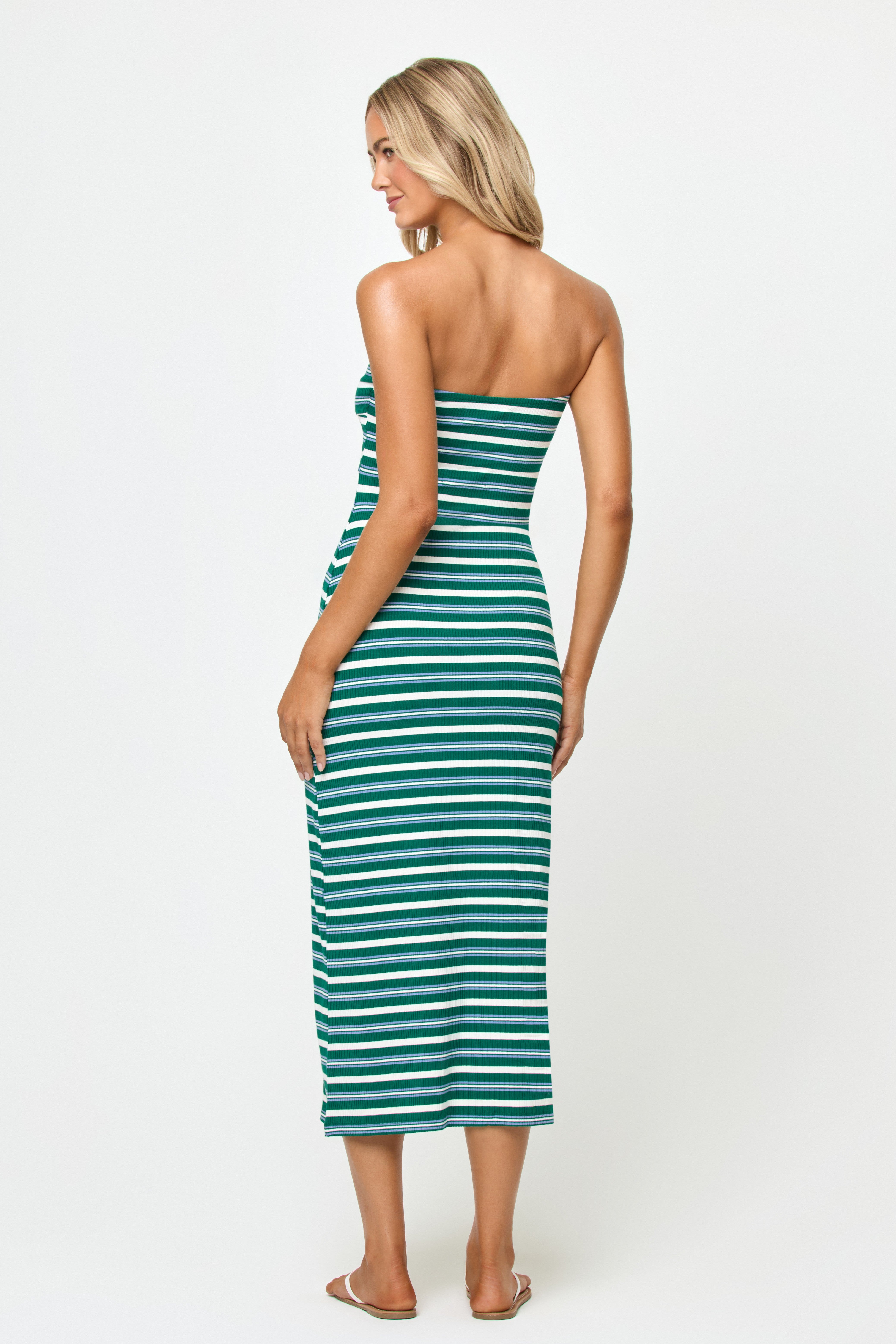 A woman with long blonde hair wears the LSpace Lisette Midi Dress—a strapless, ankle-length green and white striped dress with a twist front—and clear sandals. She stands facing away from the camera against a plain white backdrop.