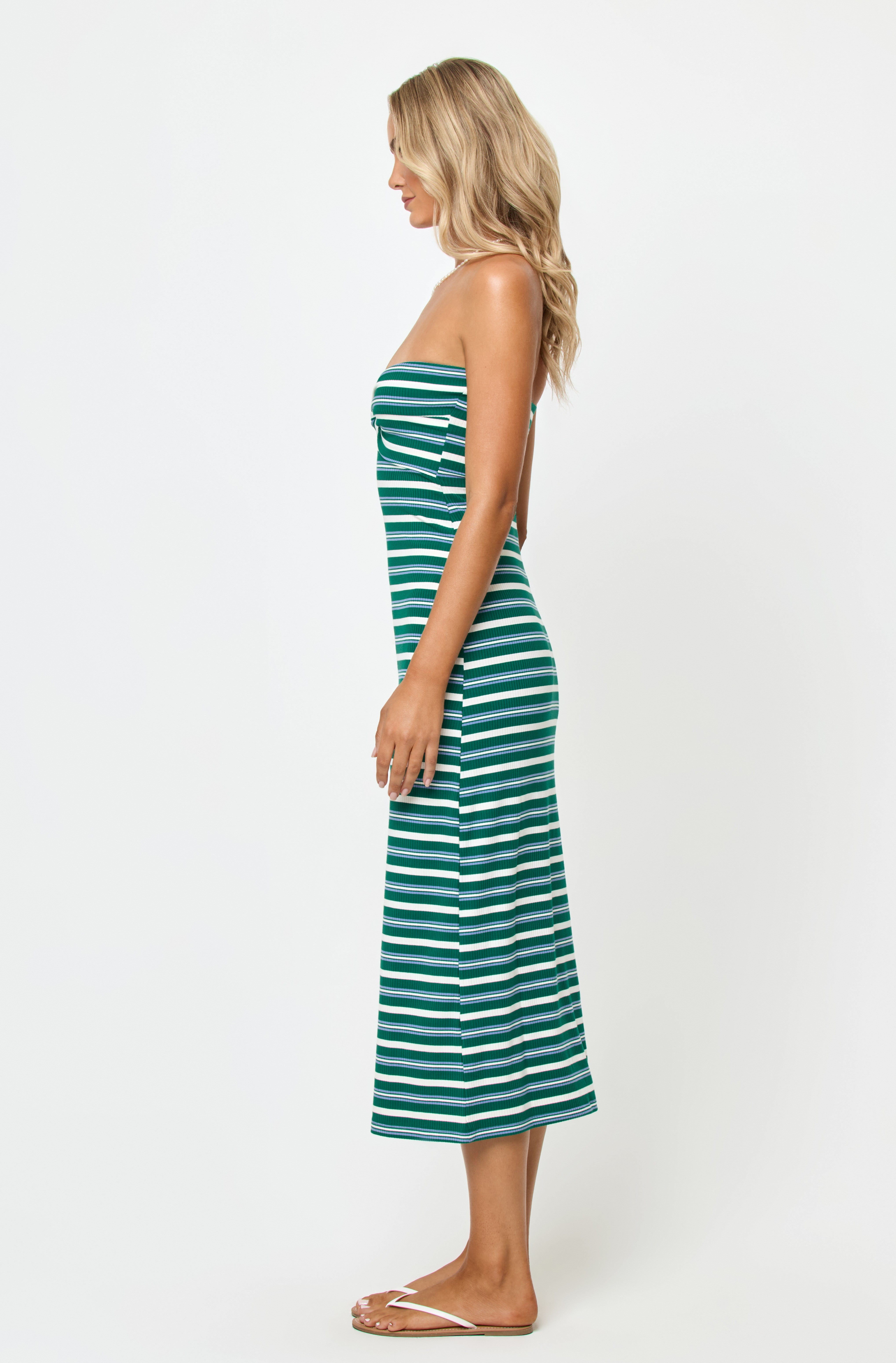 A woman in profile stands against a white background wearing the LSpace Lisette Midi Dress, featuring green and white horizontal stripes, a strapless twist front detail, and white sandals. Her wavy blonde hair completes the look.