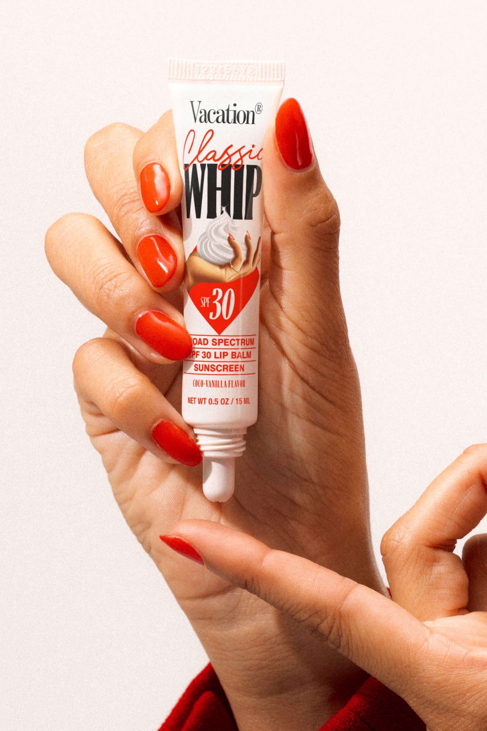 A hand with red-painted nails holds a Vacation Class Whip SPF 30 Lip Balm, while another hand points to the lightweight, water-resistant tube.