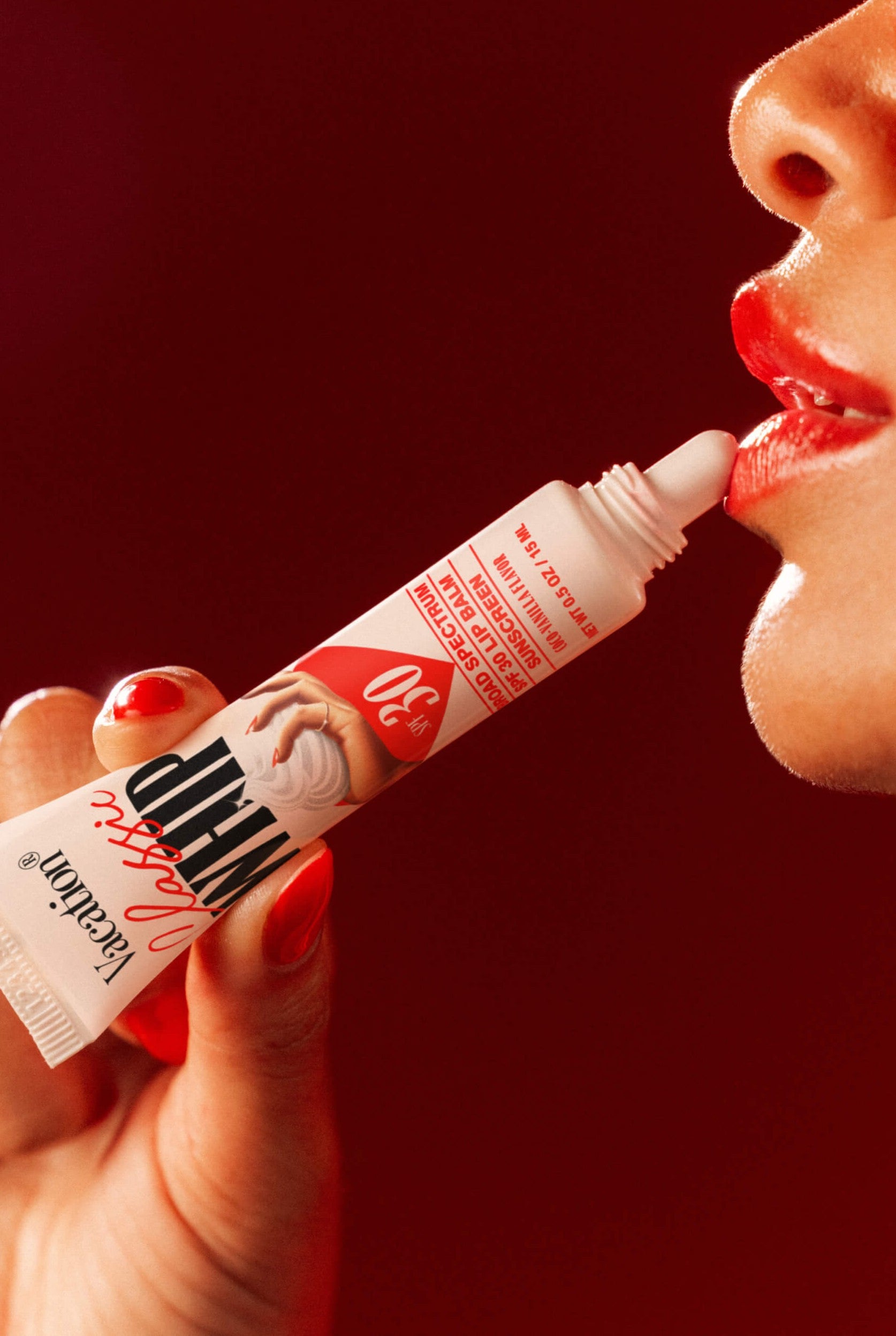 A person applies Vacation's Class Whip SPF 30 Lip Balm from a small tube to their red lips, with a deep red background and dramatic lighting emphasizing their mouth and hand.