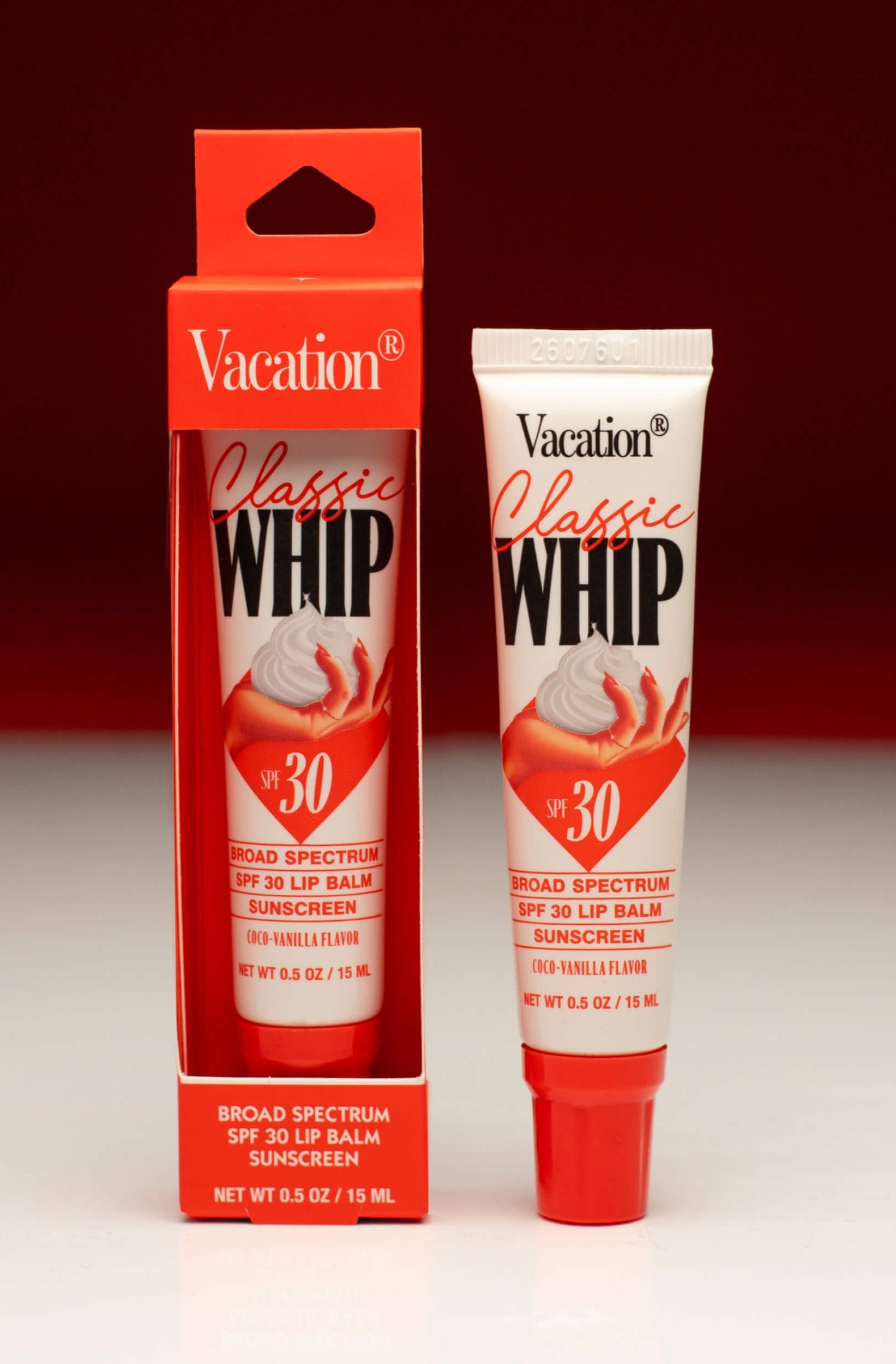 A Vacation Class Whip SPF 30 Lip Balm in Red Multi is shown with its box against a red and white background, both displaying red, white, and black design elements.