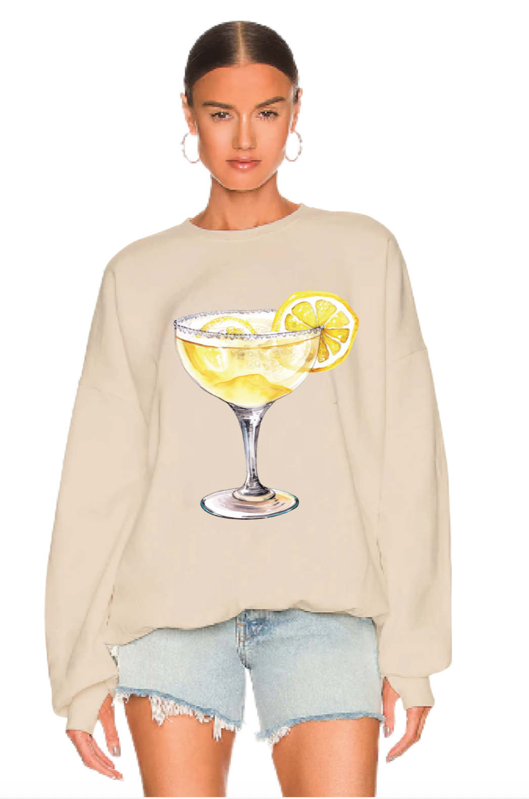 A woman wears The Laundry Room's beige "When Life Gives You Lemons Jumper," an oversized cotton-polyester sweatshirt with a large margarita glass and lemon graphic, styled with light blue denim shorts and hoop earrings.