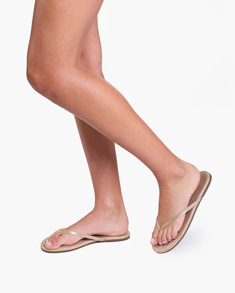 A person wearing Tkees' Glitters Tkees in Pink Pearl flip-flops stands on a white background, showing only their legs and feet. The sandals have thin straps, a minimalist design, and are made in Brazil.