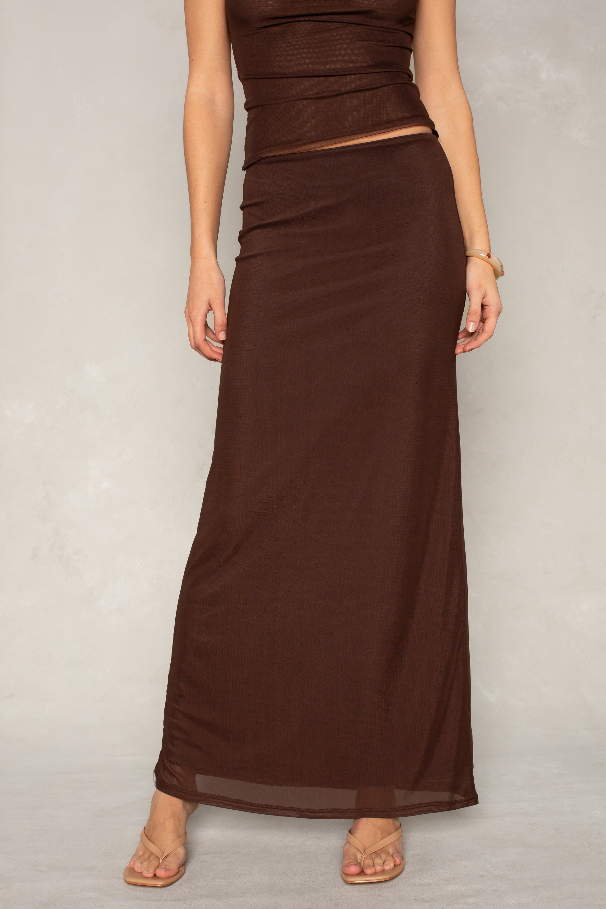 A person models the Rachael Mesh Midi Skirt by Isla x Tate, paired with a sleeveless brown fitted dress and tan sandals against a light gray background. Their head is not shown in the photo.