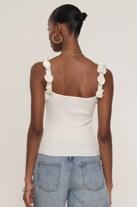 A woman is seen from behind wearing the Heartloom Marone Top, a white ribbed-knit piece with floral strap details, paired with blue jeans. Her hair is in a neat bun, complemented by gold hoop earrings and bracelets.