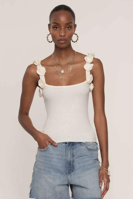 A woman wears the Marone Top by Heartloom in ivory—a ribbed-knit style with flower straps—paired with layered gold necklaces, hoop earrings, and light blue jeans. She stands against a plain background, looking at the camera with one hand in her pocket.