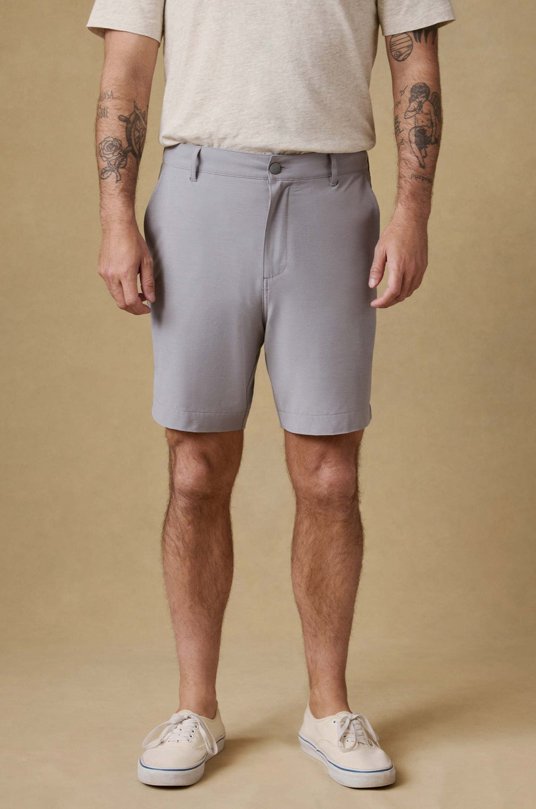 A person wearing a beige t-shirt, Faherty All Day Short 7 In in light gray, and beige sneakers stands against a tan background. Their tattooed arms are visible from the elbows down; their head is not shown.