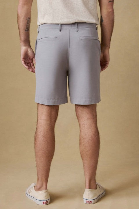 A person stands facing away, wearing a light beige t-shirt, Faherty All Day Short 7 In in light gray, and beige slip-on shoes. The plain, light tan background highlights the visible tattoos on their arms and legs.