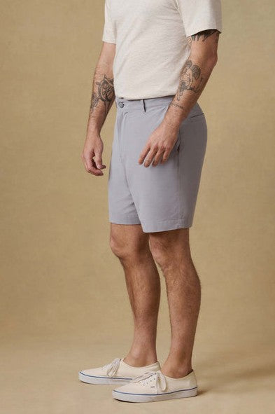 A man with tattooed arms stands sideways against a beige background, wearing a white short-sleeve shirt, Faherty All Day Short 7 In in light gray, and cream sneakers.