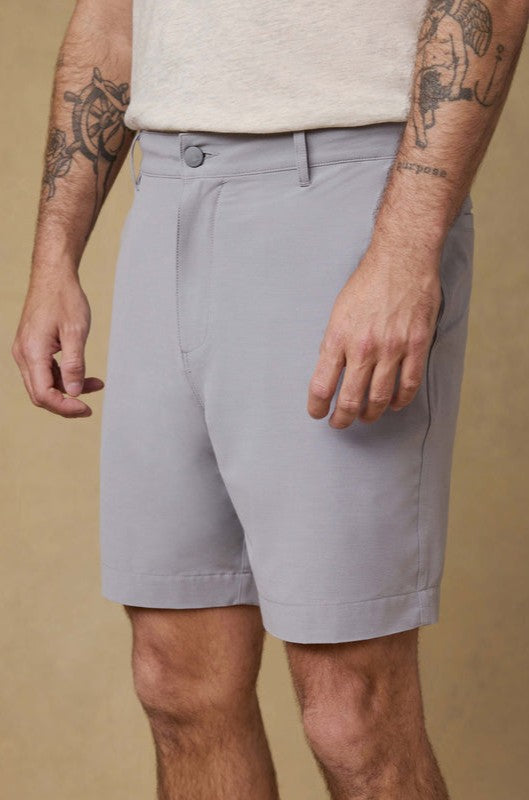 Wearing Faherty’s All Day Short 7 In in light gray and a beige tee, a person with arm tattoos stands against a tan backdrop. The image is cropped from shoulders to mid-thigh.