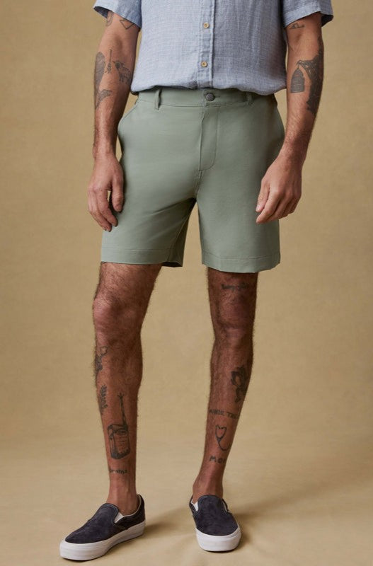 Against a beige background, a person (face not visible) wears a light blue short-sleeve button-up shirt and Faherty All Day Short 7 In in sage green with black slip-on shoes. Their tattooed arms and legs are visible.
