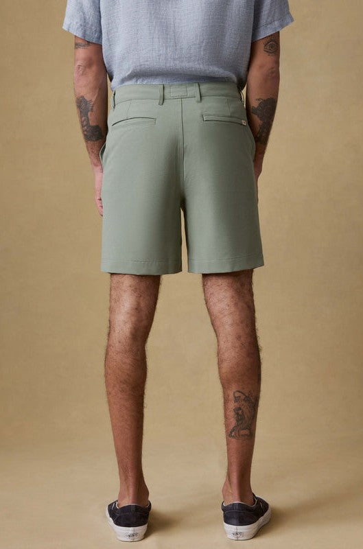 A person faces away from the camera, wearing Faherty's All Day Short 7 In in light green organic cotton and a light gray shirt. Their tattooed legs and dark slip-on shoes are visible against a neutral beige background.