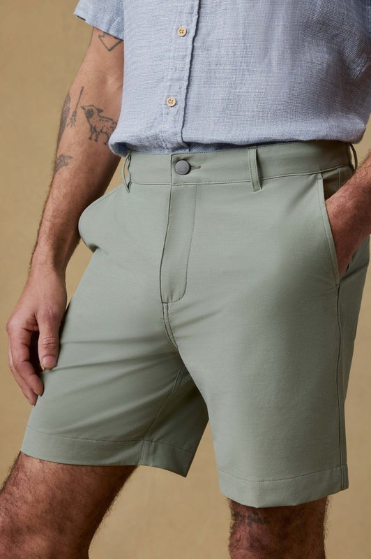 A person wears Faherty All Day Short 7 In in light green with side pockets and a light blue short-sleeve button-up. Their left hand is in their pocket, displaying arm tattoos, against a beige background for a neutral vibe.