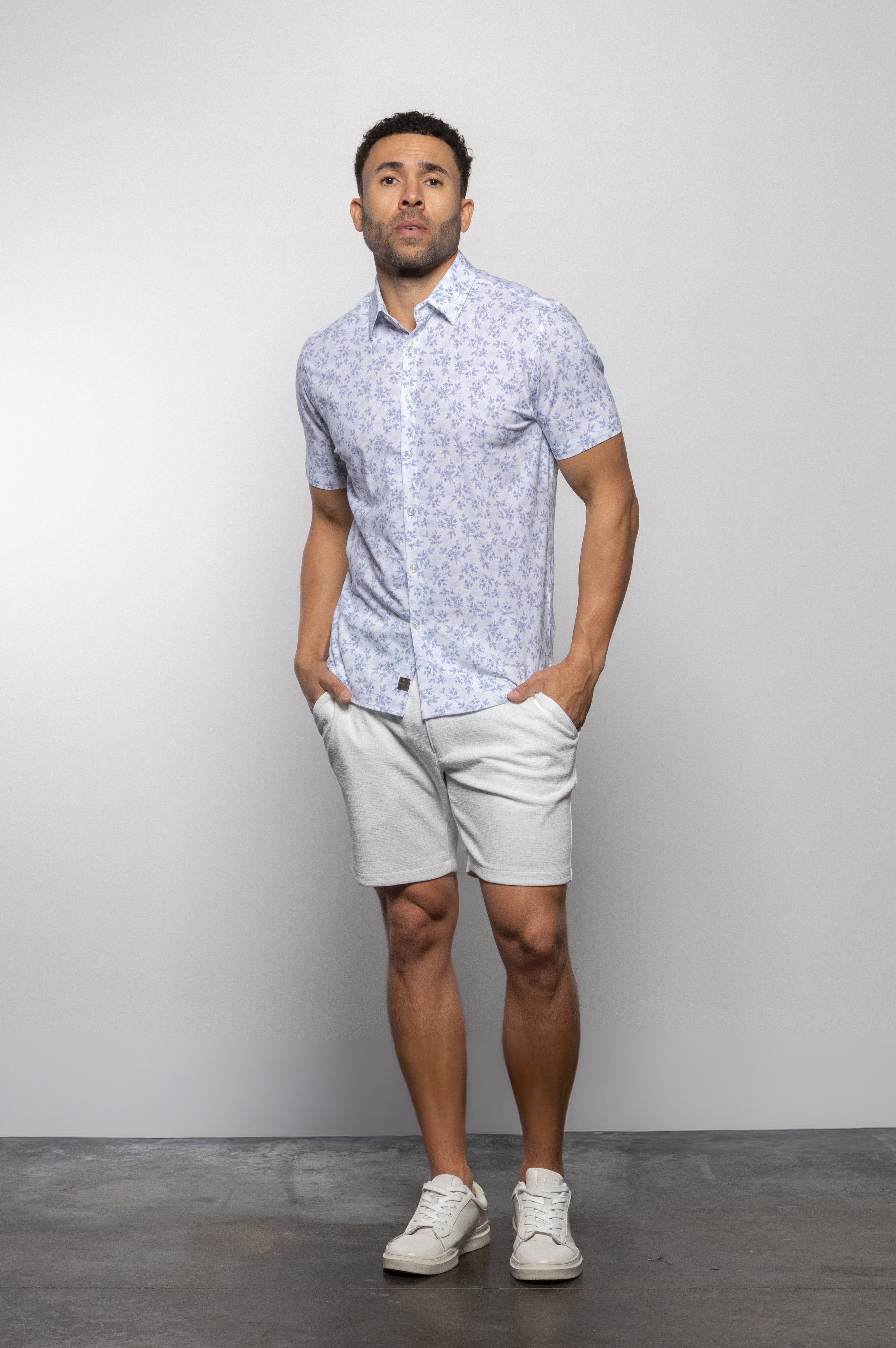 A man stands against a plain background, wearing Mason Cruz's Bello Wildflower light blue patterned short-sleeve tailored fit shirt with a casual collar, paired with light gray shorts and white sneakers, his hands in his pockets.
