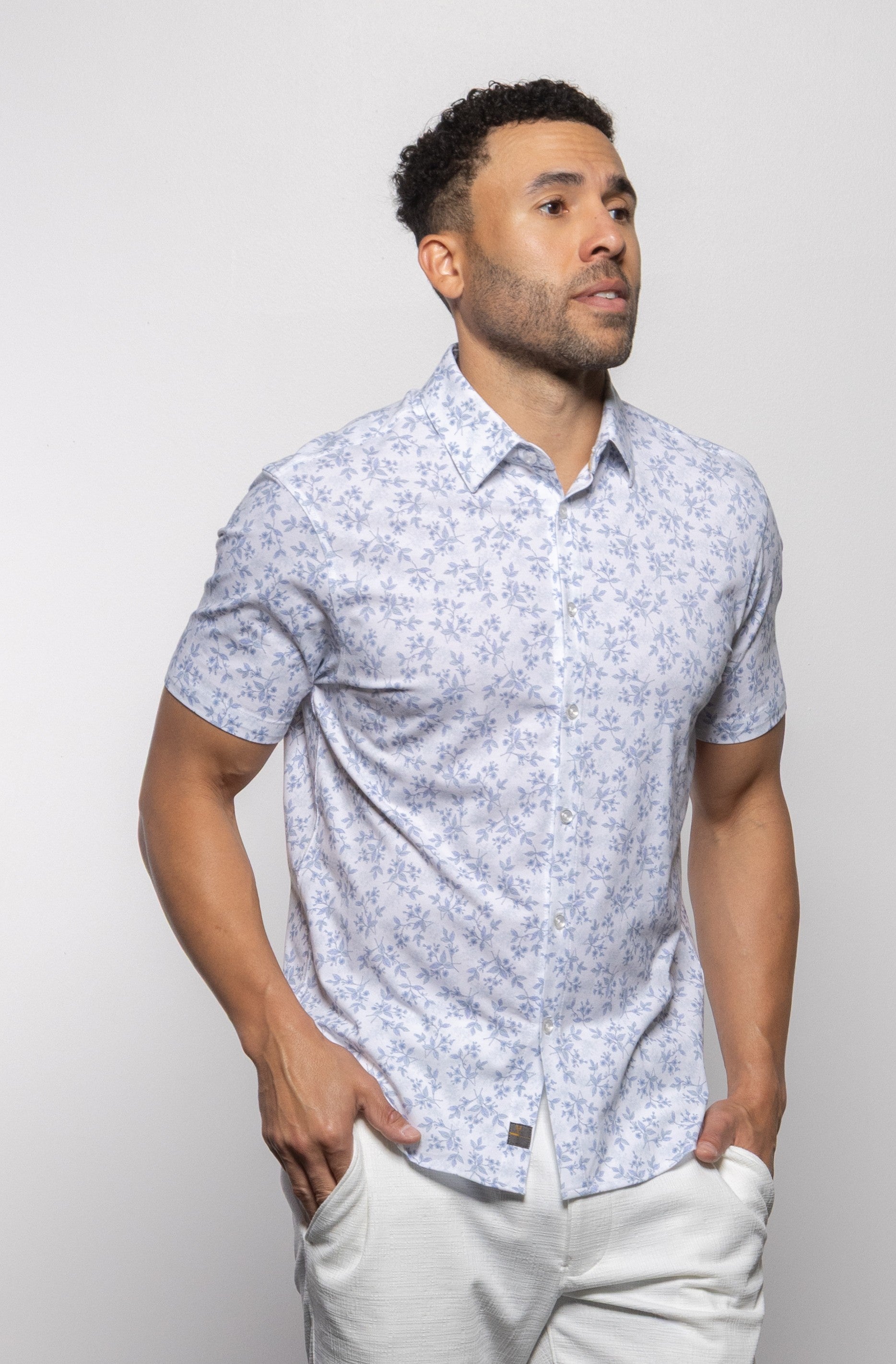 A man wearing the Mason Cruz "Bello Wildflower" tailored fit shirt in SUMMER WHITE, featuring a light blue floral print, short sleeves, a casual collar, and paired with white pants, stands with his hands in his pockets against a plain white background.