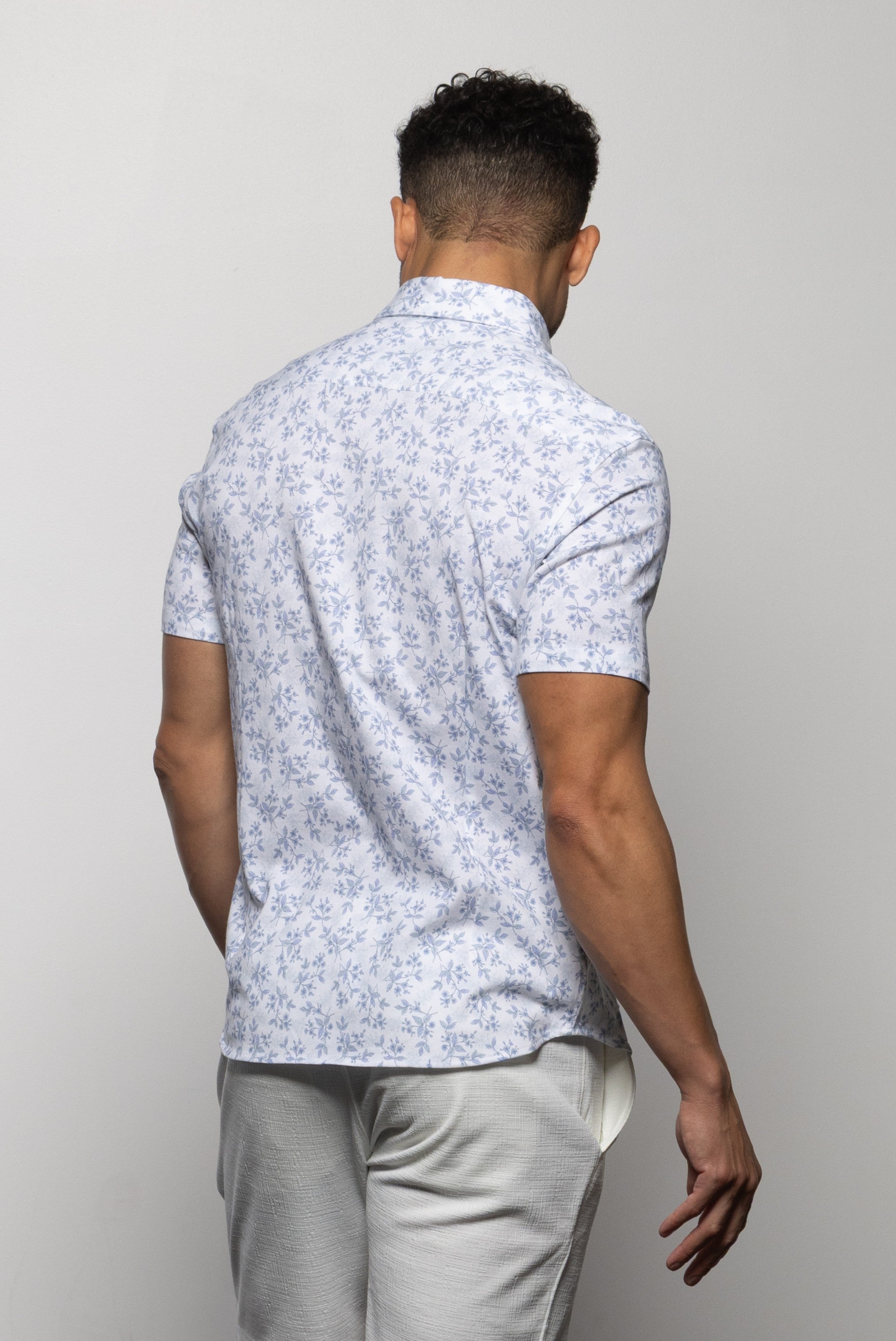A man stands facing away, wearing the Mason Cruz "Bello Wildflower" light blue floral short-sleeve tailored fit shirt with a casual collar and light-colored pants against a plain background.