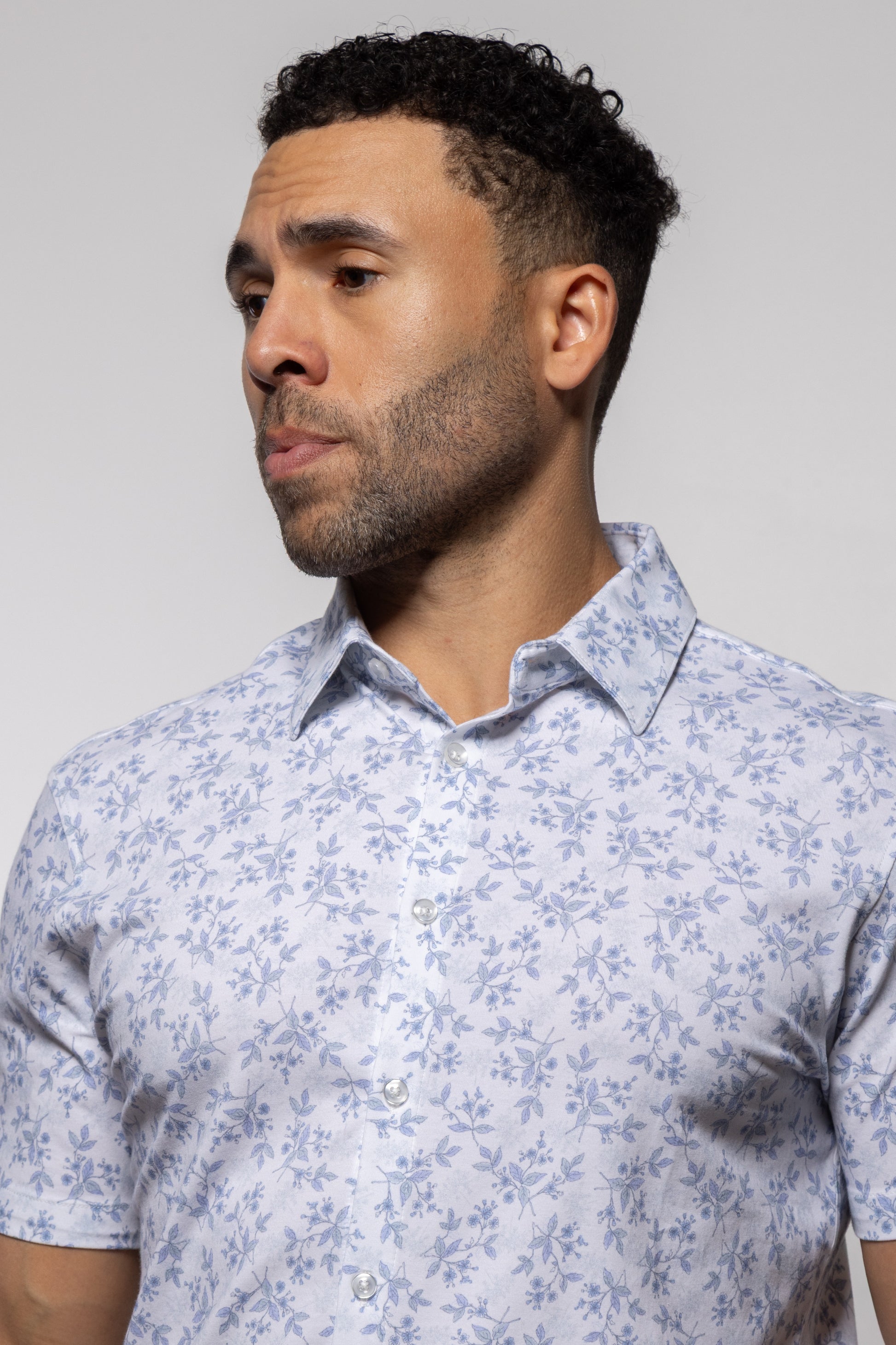 A man with short curly hair wearing the Mason Cruz Bello Wildflower light blue floral short-sleeve shirt with a casual collar looks to the side against a plain background.