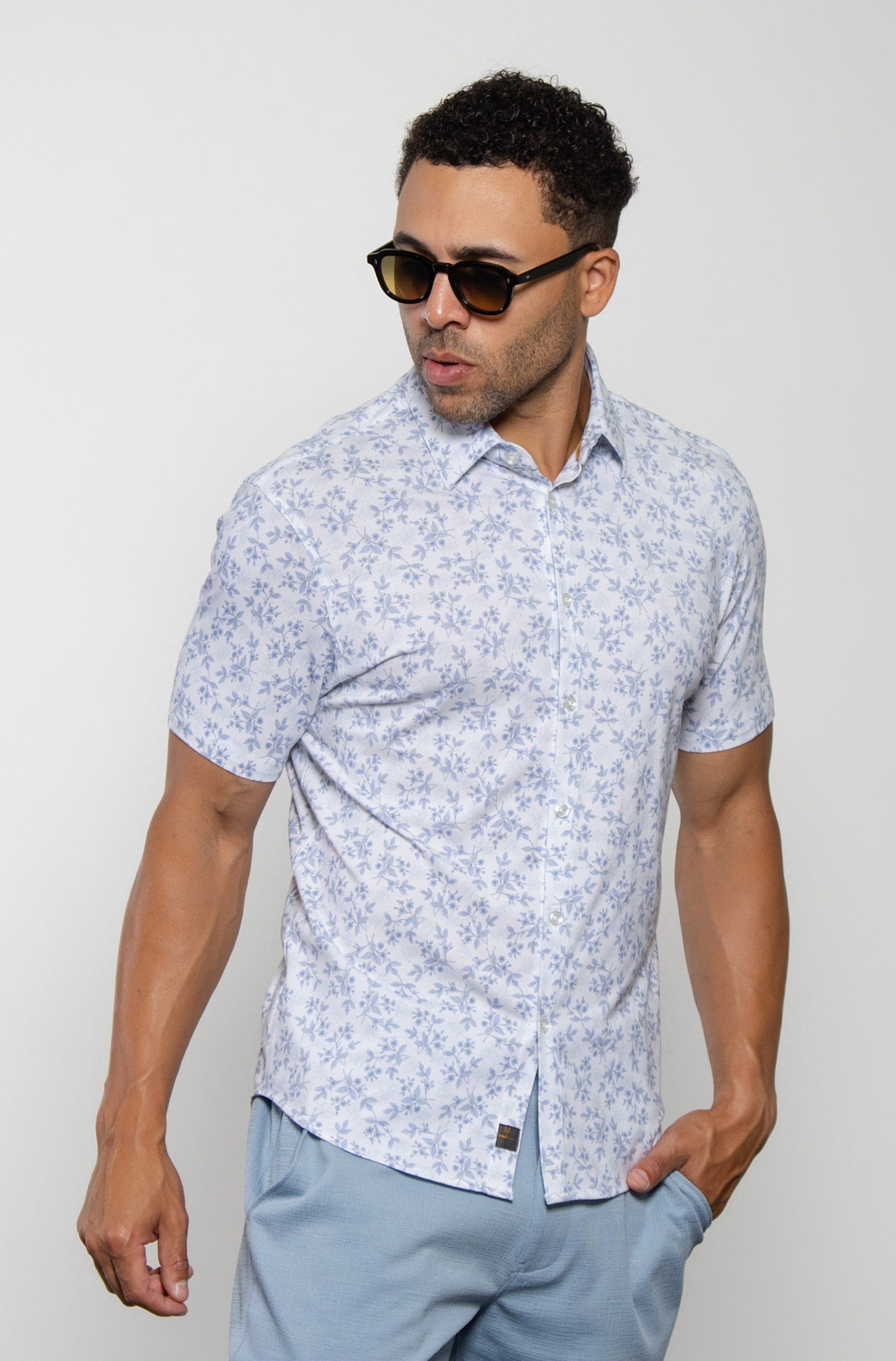 A man wearing sunglasses and light blue pants stands against a plain white background with one hand in his pocket, looking off to the side. He is dressed in the Mason Cruz "Bello Wildflower"—a light blue floral short-sleeve tailored fit shirt featuring a casual collar and crafted from premium cotton blend.