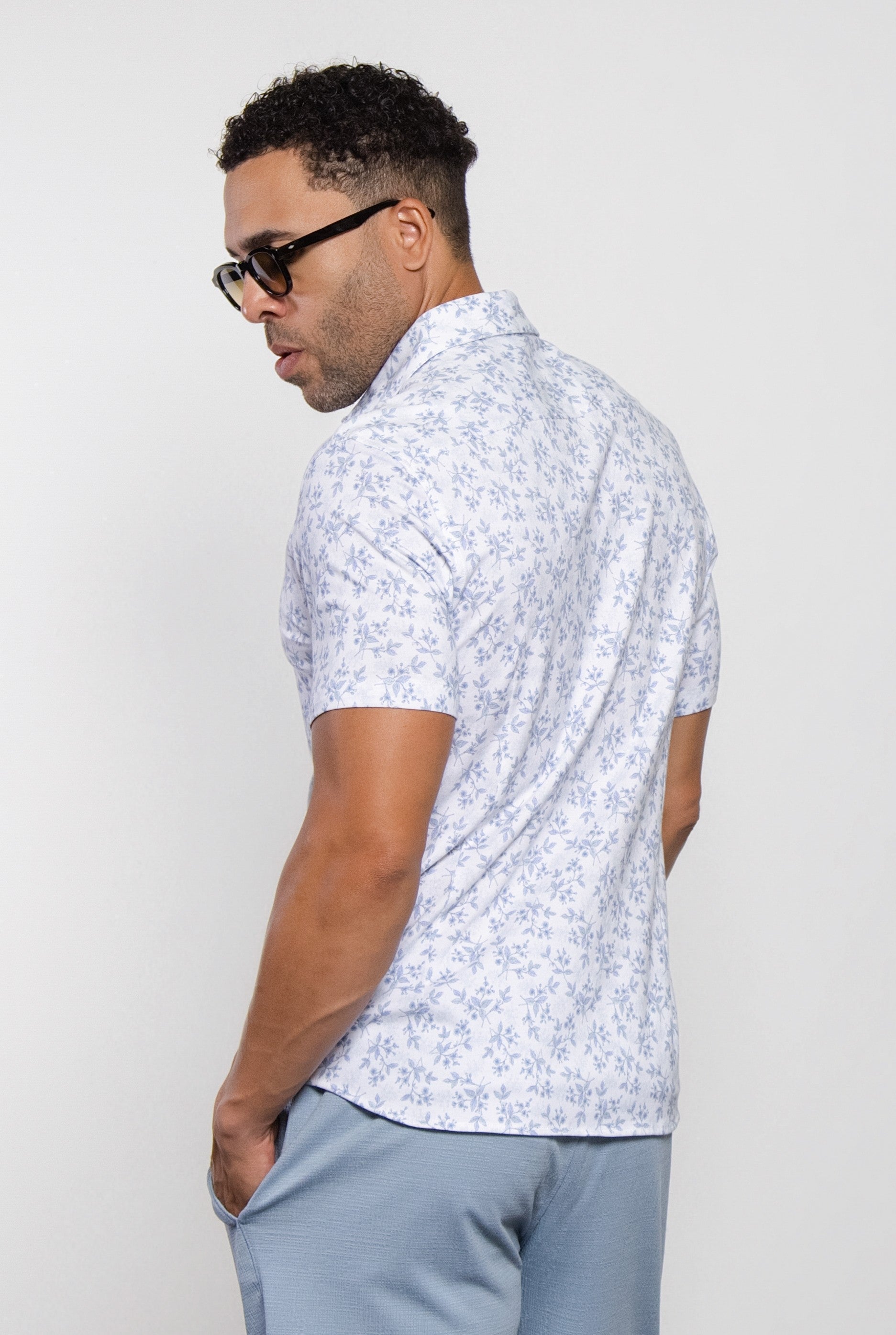 A man wearing the Mason Cruz "Bello Wildflower" light blue floral short-sleeved tailored fit shirt with a casual collar and sunglasses stands with his back to the camera, one hand in his pocket, posing against a plain white background.