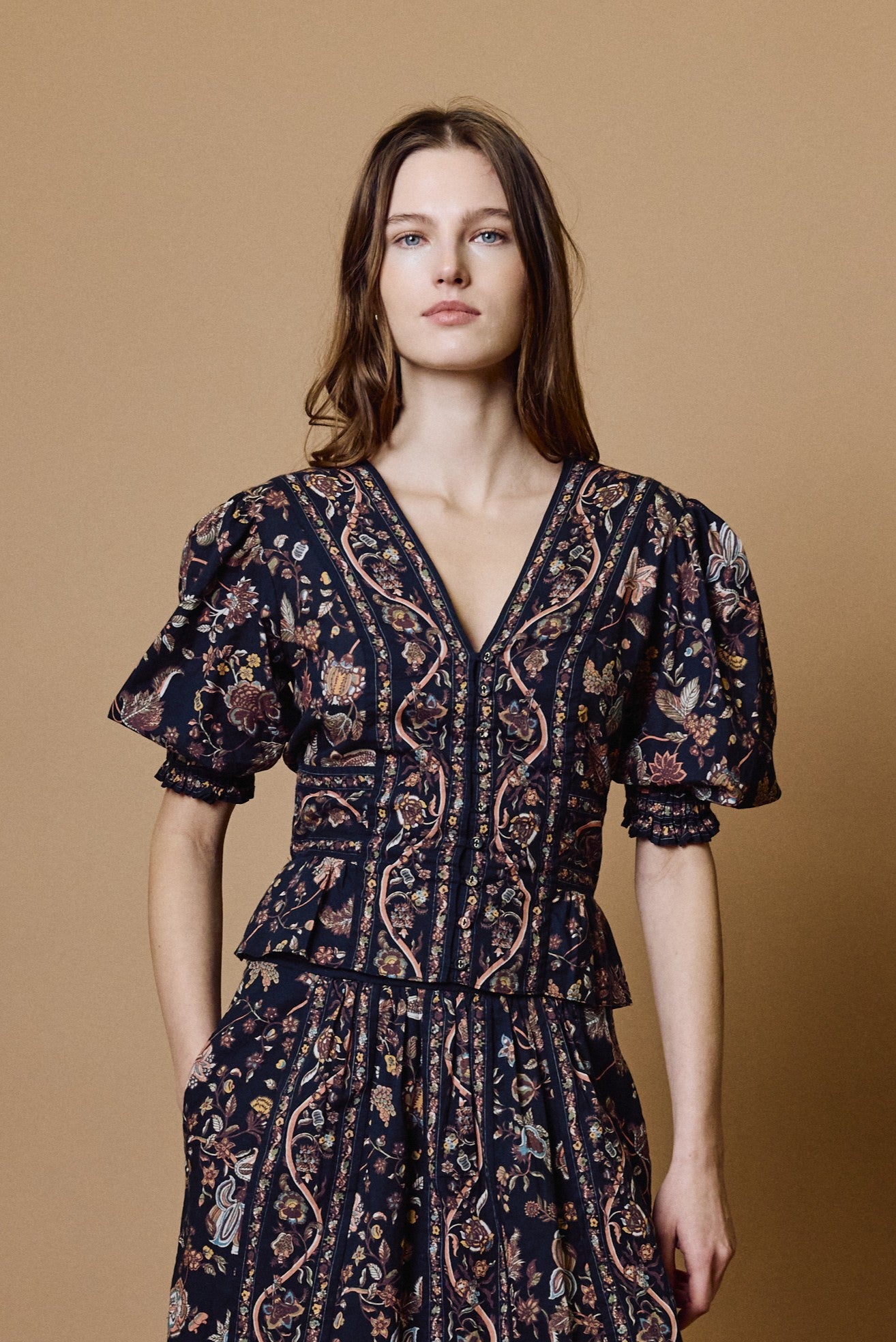 A woman with long brown hair wears the Melissa Top by Cleobella in the BOIS PRINT BOIS color, featuring a dark floral pattern, short puffed sleeves, and a V-neckline as she stands against a plain beige background. The elegant puffed sleeves of this chic Melissa Top add a romantic touch to its floral design, creating a timeless look.
