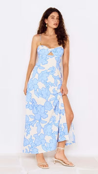 A woman with long curly hair wears the Minkpink Alessia Twist Front Midi Dress, featuring a blue floral print, twist-front cutout, spaghetti straps, and high slit—styled with strappy sandals and hoop earrings for summer events.