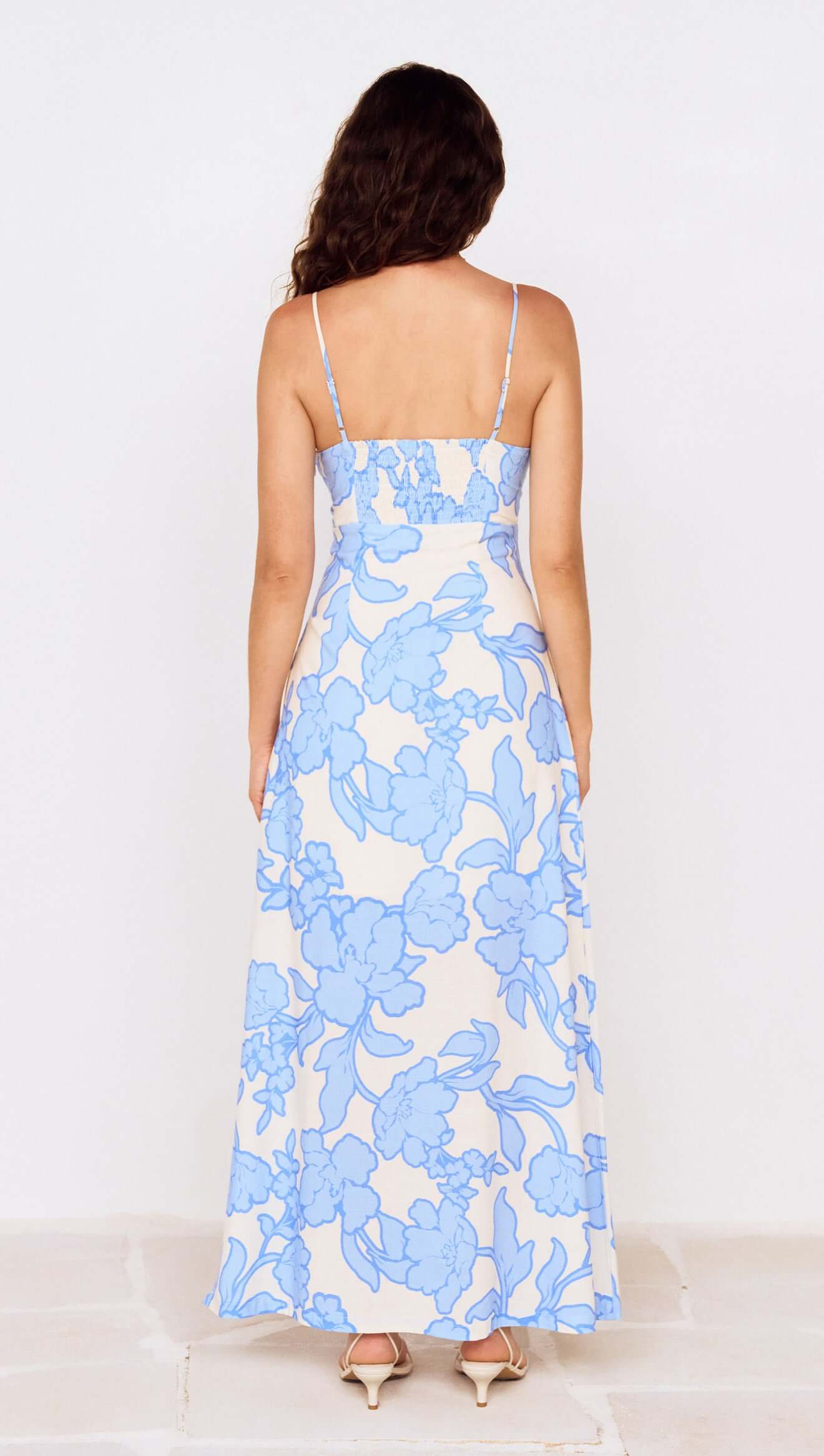 A woman with wavy brown hair wears the Minkpink Alessia Twist Front Midi Dress, featuring a blue floral print on a white base and thin straps. She stands with her back to the camera, highlighting this perfect summer event dress.