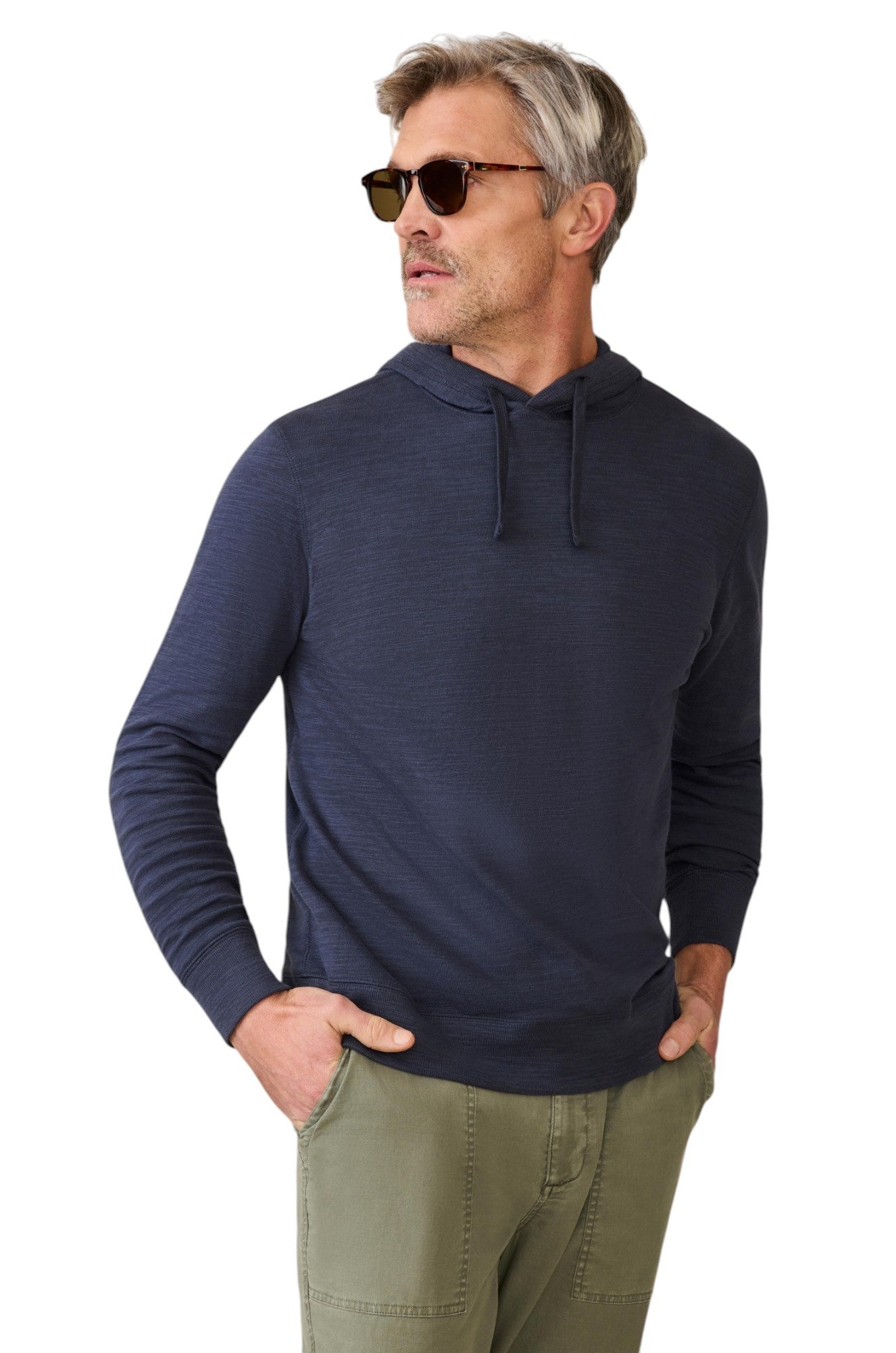 A man in sunglasses and olive green pants stands with his hands in his pockets against a white background, wearing the Faherty Sunwashed Slub Hoodie in dark blue.