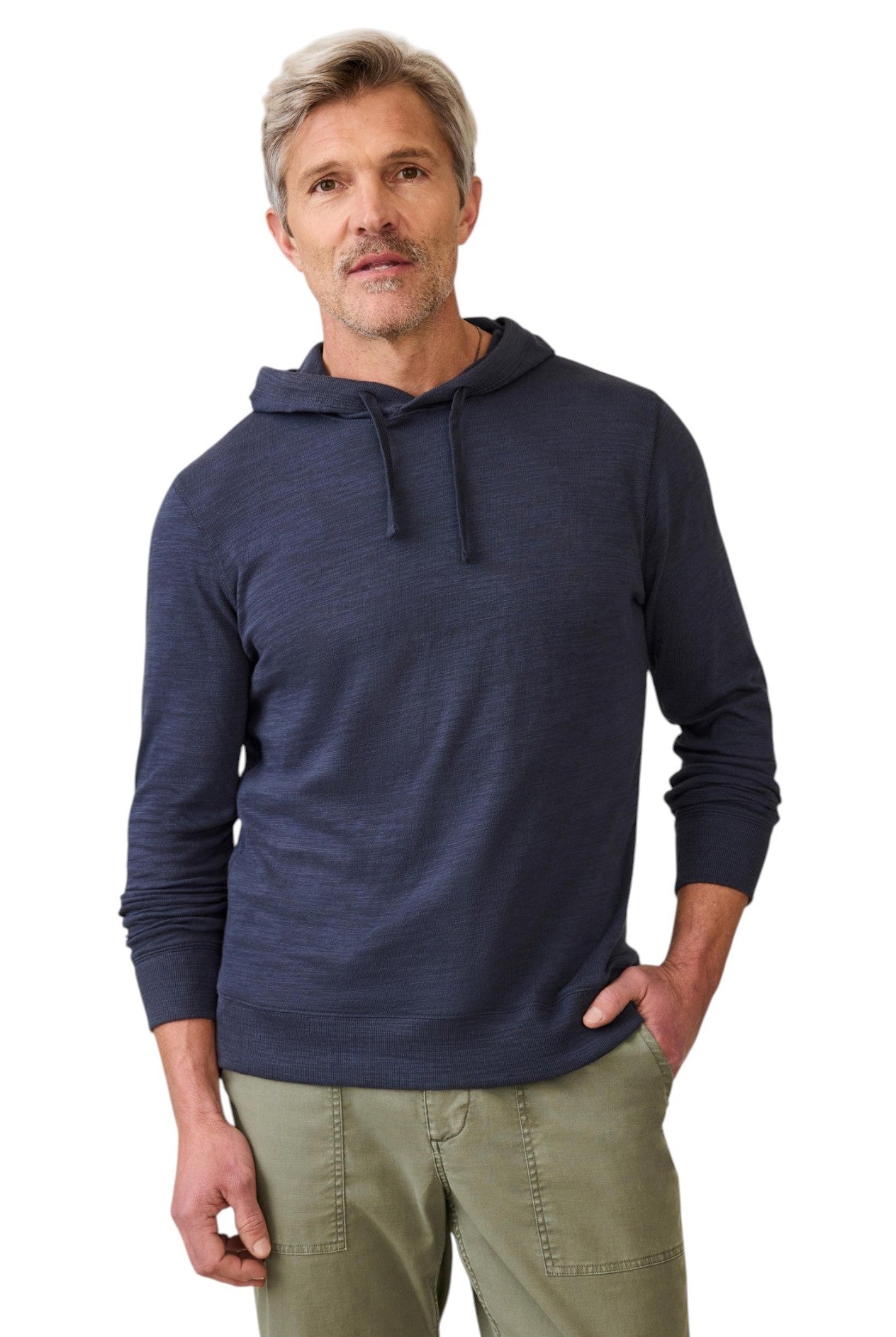 A middle-aged man with short gray hair and a trimmed beard wears the Faherty Sunwashed Slub Hoodie in Navy and olive green pants, standing with one hand in his pocket against a white background.