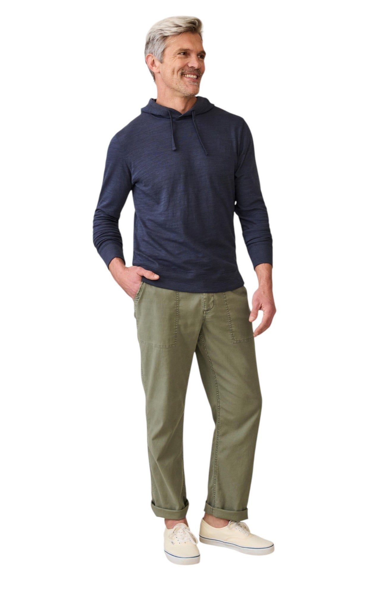 A smiling man with gray hair wears the Faherty Sunwashed Slub Hoodie in navy blue, olive green pants, and beige sneakers. He stands casually with one hand in his pocket against a white background.