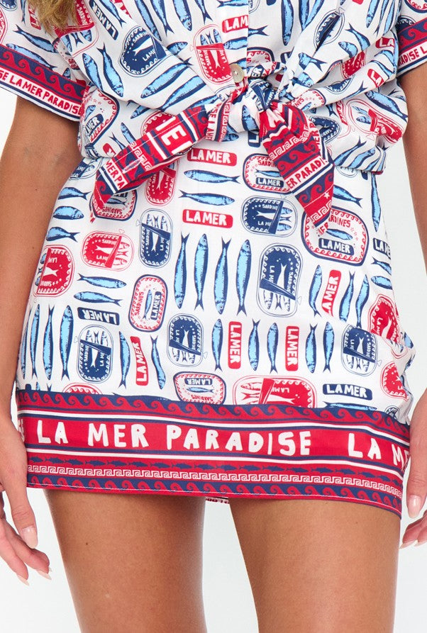 A close-up of a woman’s lower torso wearing the All Night Skort by Show Me Your Mumu. This 100% cotton skort features red, blue, and white patterns with phrases like LA MER, PARADISE, and motifs of fish and cans.