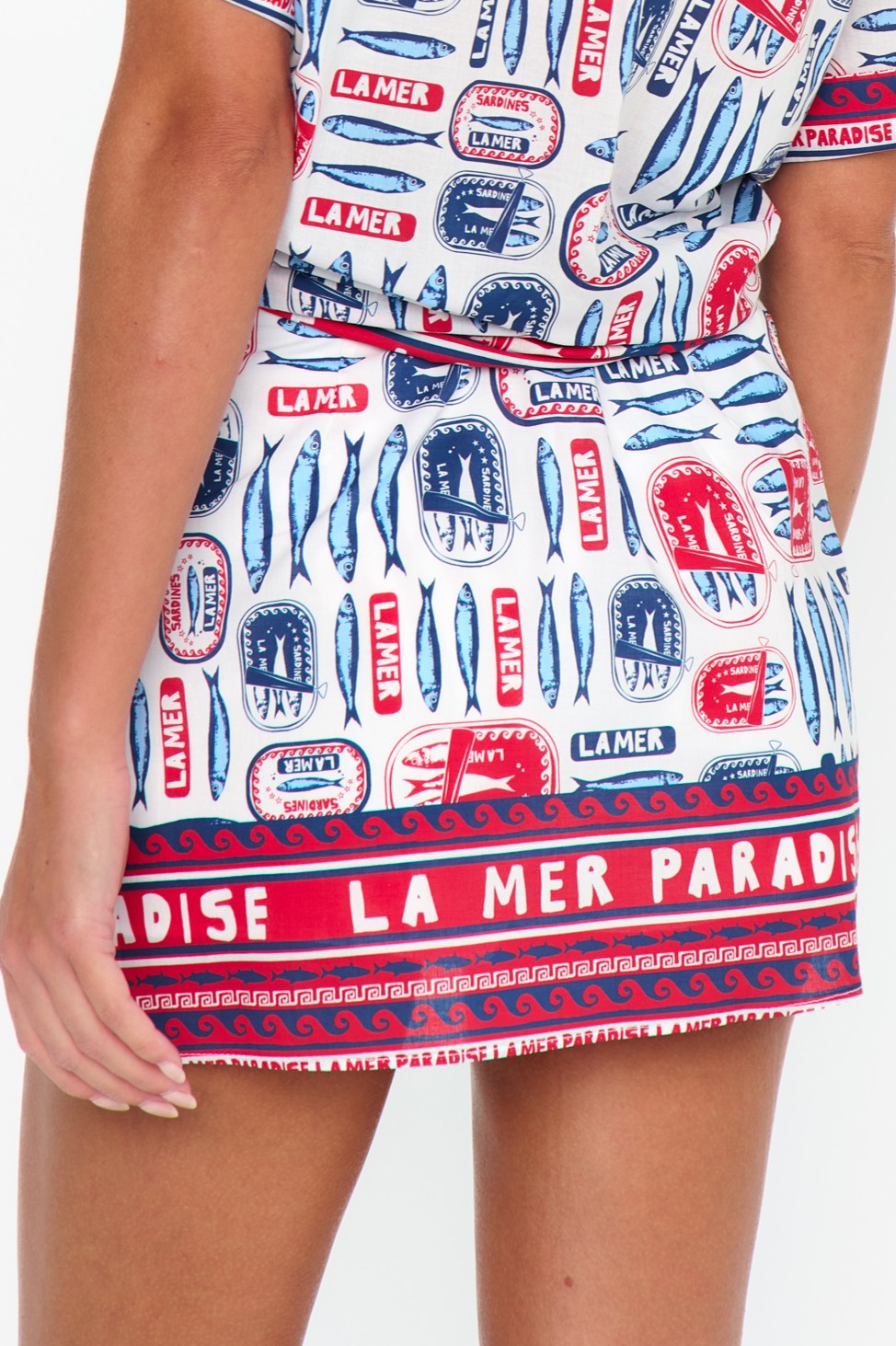 A person wears the Show Me Your Mumu All Night Skort, a colorful 100% cotton piece with red, blue, and white fish patterns and "LA MER" and "PARADISE" repeated along the hem. Their hand rests by their side.