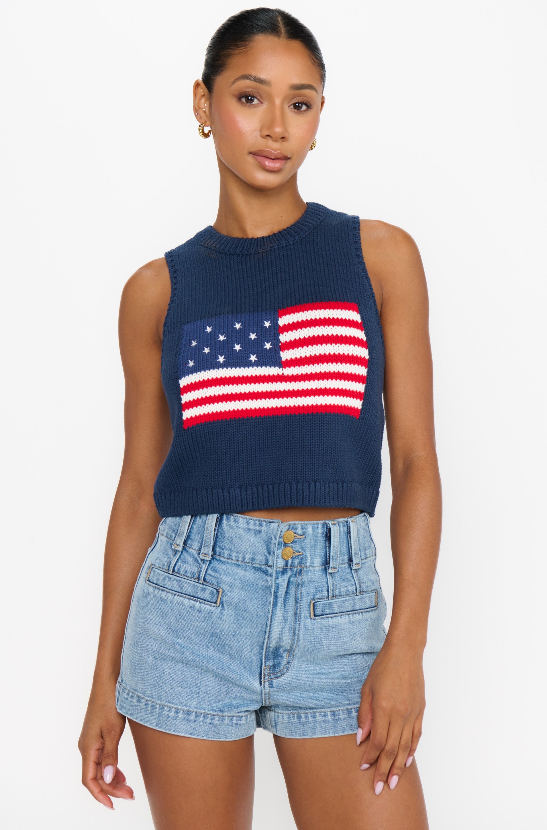 A woman wears the Show Me Your Mumu Milo Tank in Blue Multi—a relaxed fit, sleeveless knit top with an American flag design—paired with high-waisted light blue denim shorts, standing against a plain white background.