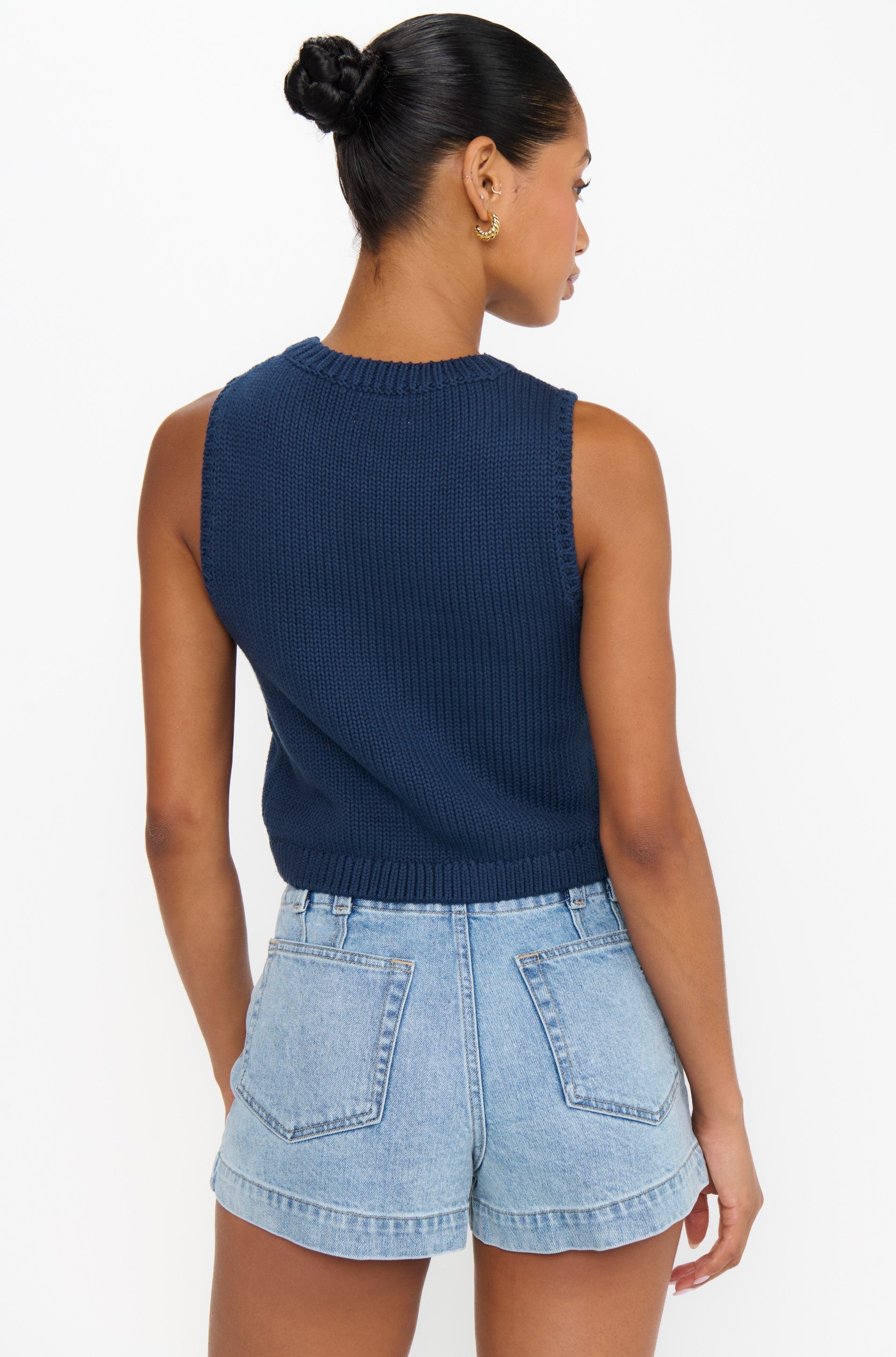 A woman with a bun is shown from the back wearing the Show Me Your Mumu Milo Tank, a sleeveless navy blue 100% cotton knit top, paired with light blue high-waisted relaxed fit denim shorts against a white background.