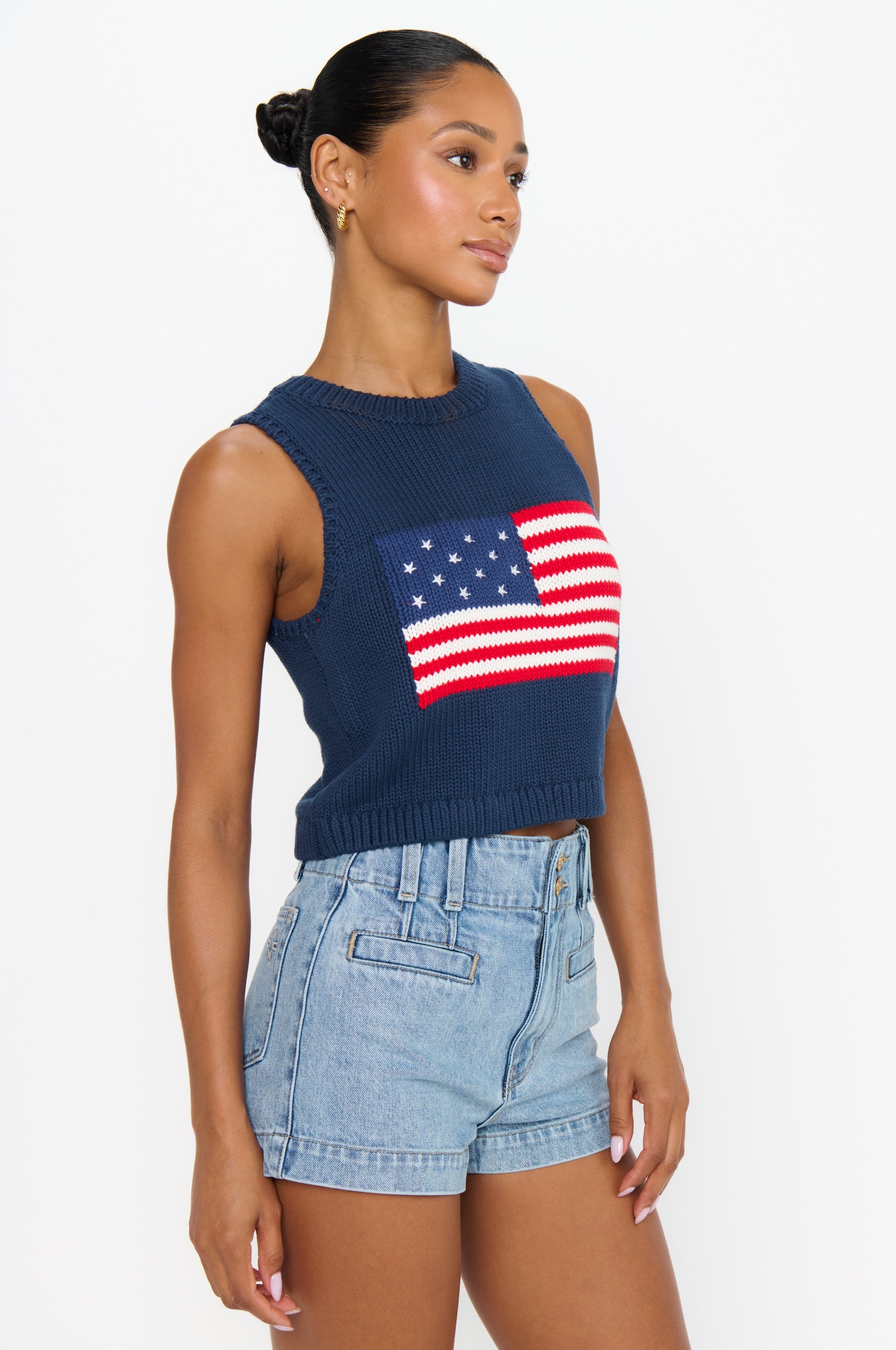 A woman wears the Show Me Your Mumu Milo Tank, a relaxed fit navy sleeveless knit top with an American flag design, paired with light blue high-waisted denim shorts, standing against a plain white background.