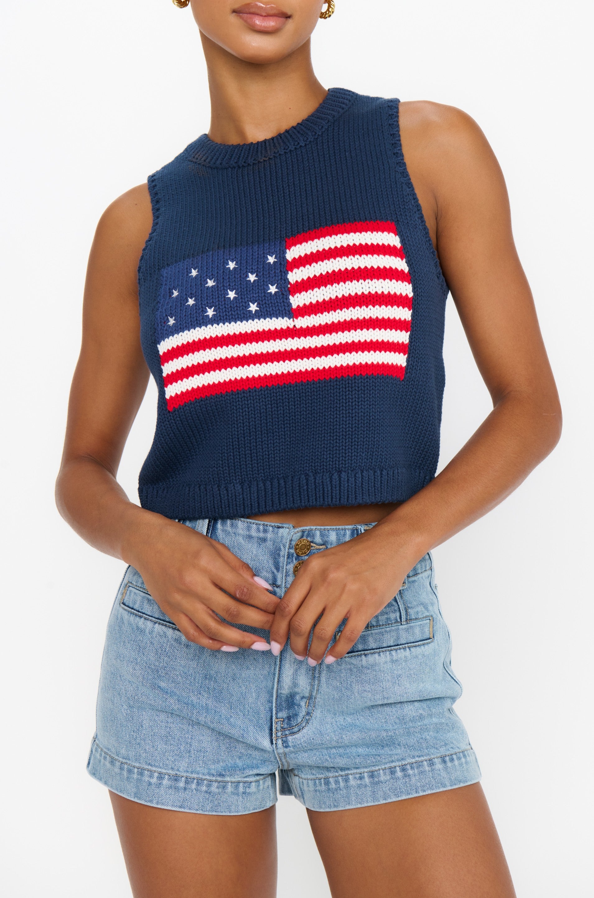 A person wears the Show Me Your Mumu Milo Tank, a relaxed fit navy sleeveless knit with crocheted American flag design, paired with high-waisted light blue denim shorts against a white background.