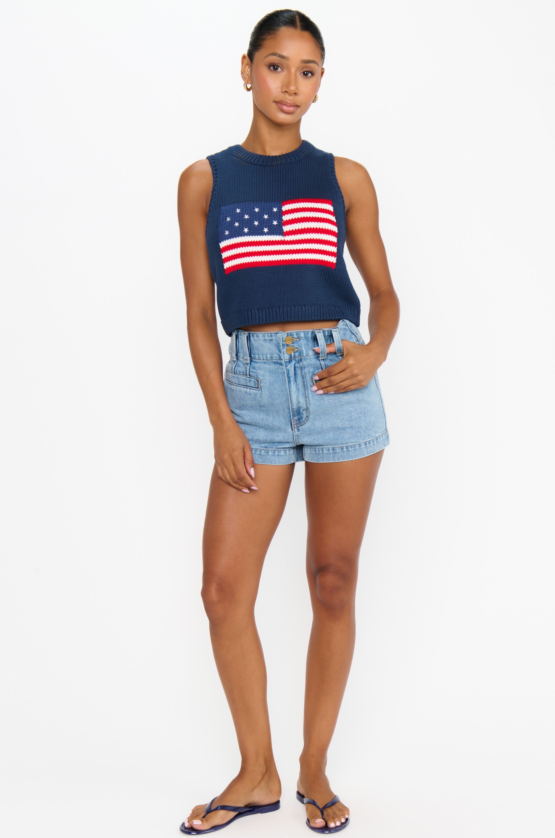 A woman stands against a white background wearing the Show Me Your Mumu Milo Tank, a navy blue 100% cotton sleeveless knit with an American flag design, light denim shorts, and dark flip-flops. Her hair is pulled back and one hand rests in her pocket.