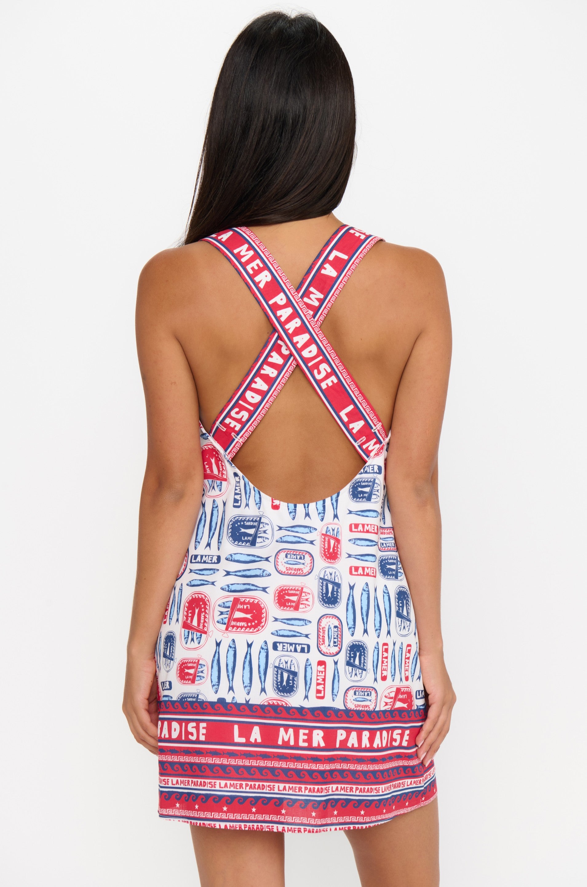 A woman with long dark hair is seen from behind wearing the Show Me Your Mumu Detour Mini Dress, a sleeveless, backless piece featuring lightweight fabric with red, blue, and white patterns and “LA MER PARADISE” on the straps and hem. White background.