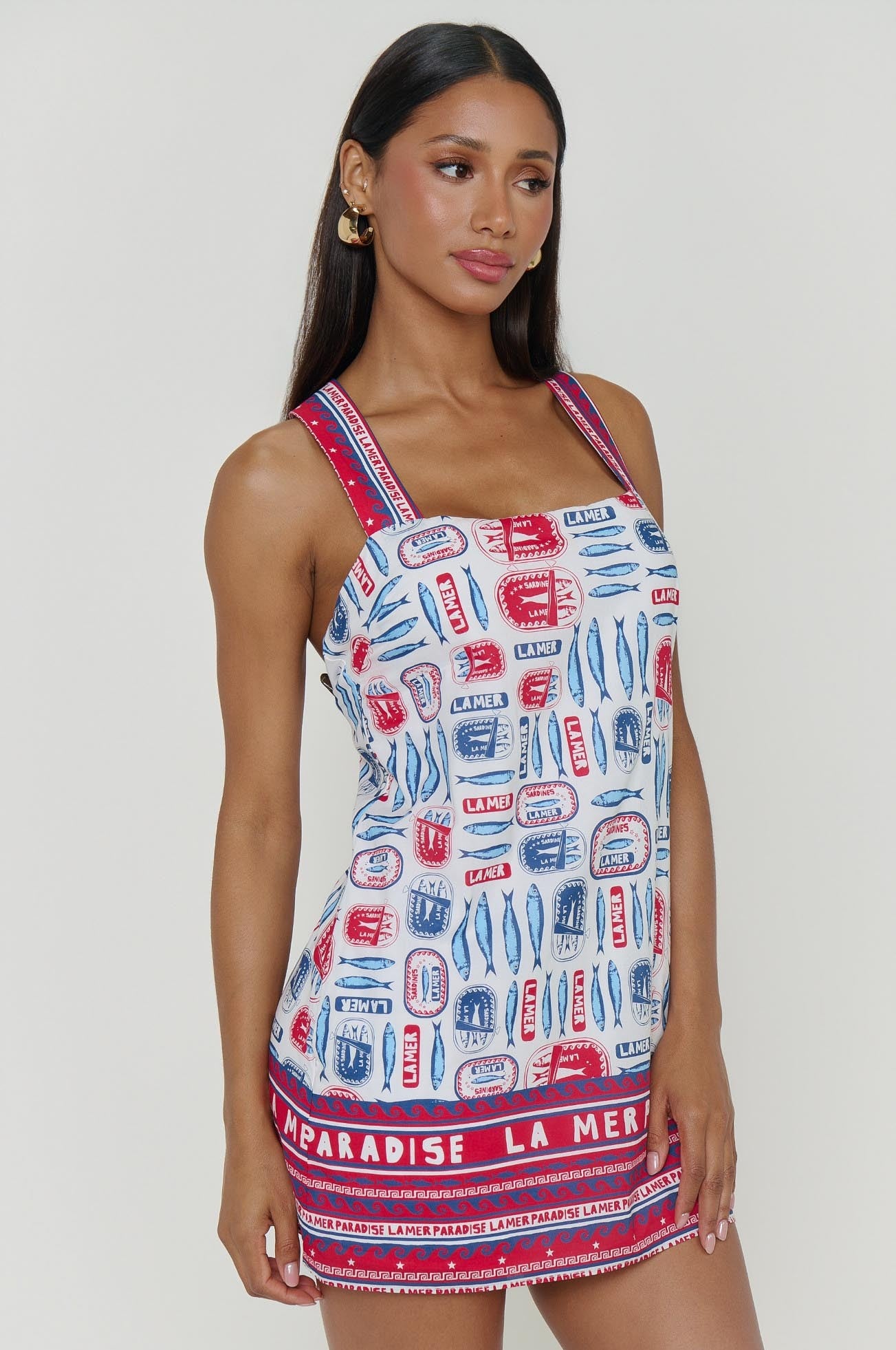 A woman stands against a white background wearing the Detour Mini Dress by Show Me Your Mumu, a colorful, sleeveless mini dress featuring red and blue sardine prints and "PARADISE LA MER" text along the hem in lightweight fabric.