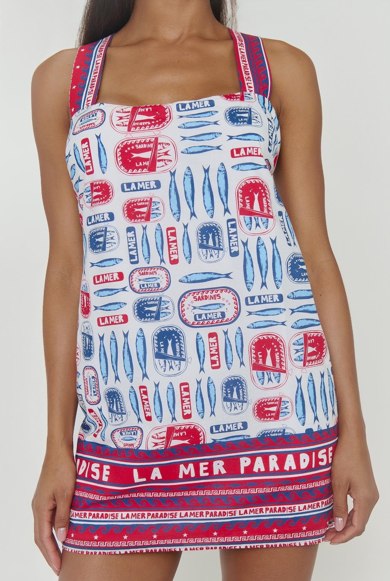 A woman wears the Detour Mini Dress by Show Me Your Mumu, a lightweight vacation dress with red, blue, and white fish and stamp patterns plus "LA MER" and "PARADISE" text on white, featuring broad straps and a square neckline.