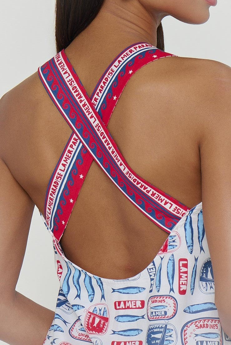 A woman wears the Detour Mini Dress by Show Me Your Mumu, featuring sleeveless sardine print with blue and red can motifs, “LA MER” text, wave patterns, and adjustable crisscross back straps—a perfect lightweight vacation dress.