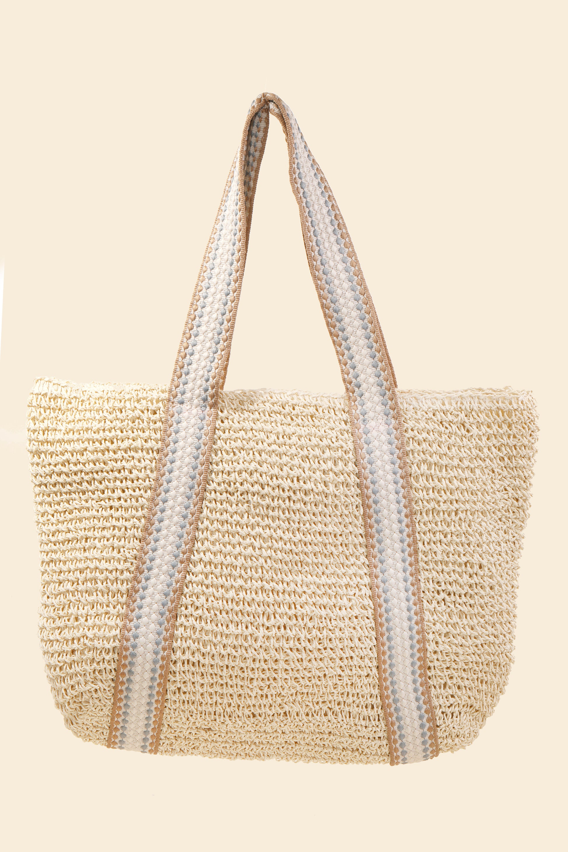 The South Moon Under Beach Tote is a woven straw bag with wide handles, featuring beige, white, and gray stripes in a classic paper style design against a light beige background.
