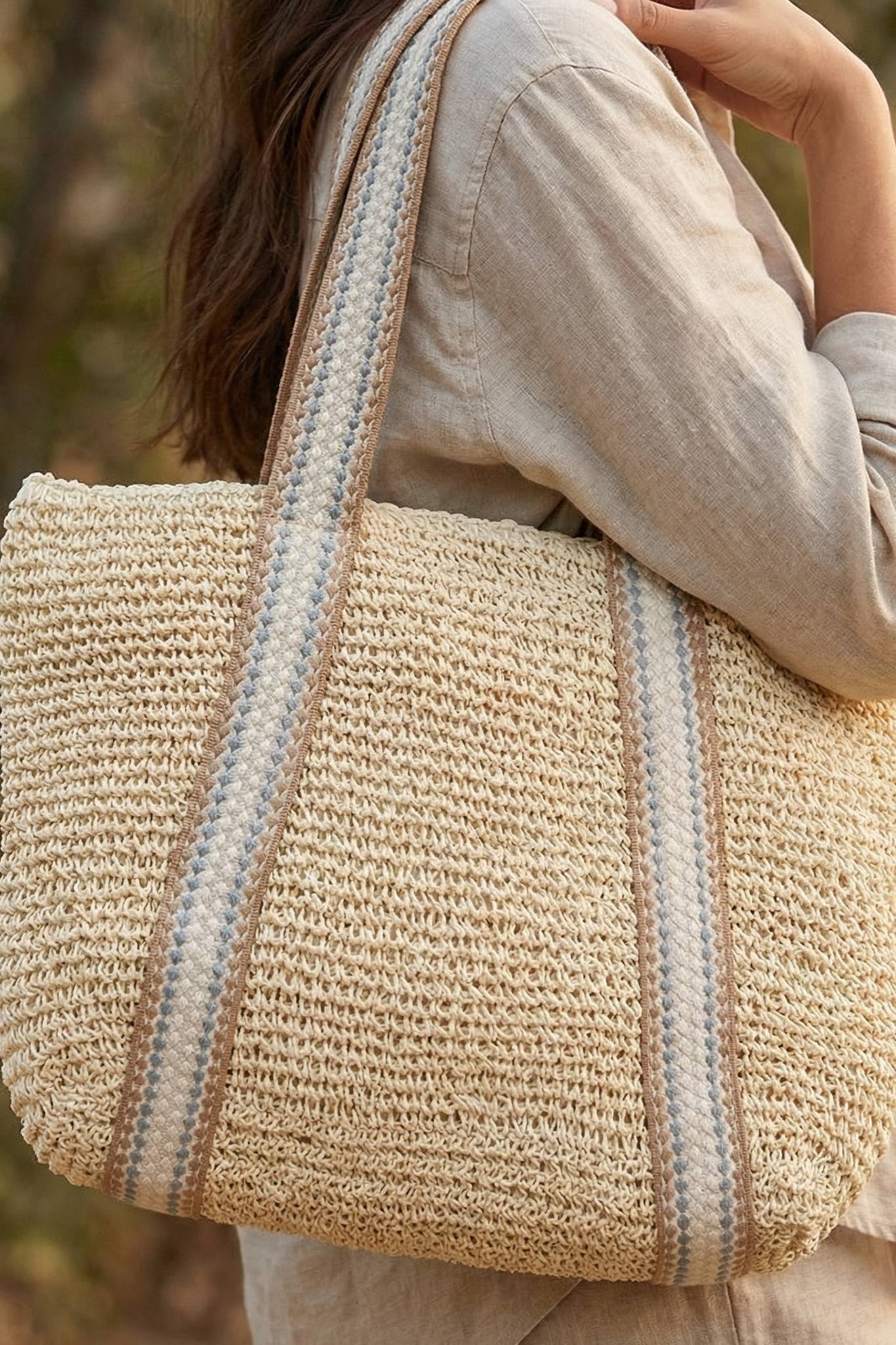 An individual in a light outfit carries the South Moon Under Beach Tote in ivory, featuring blue and beige striped straps. Outdoors with greenery, this stylish knit zipper tote is a fresh accessory for any look.