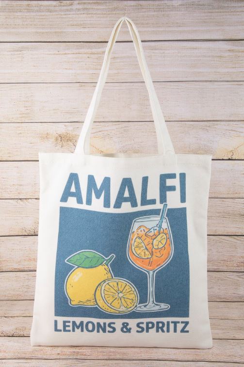 An ivory multi Canvas Tote by South Moon Under (MMA9333MU) features lemon and spritz cocktail artwork with "AMALFI LEMONS & SPRITZ" text on a blue background, shown hanging against a wooden backdrop.