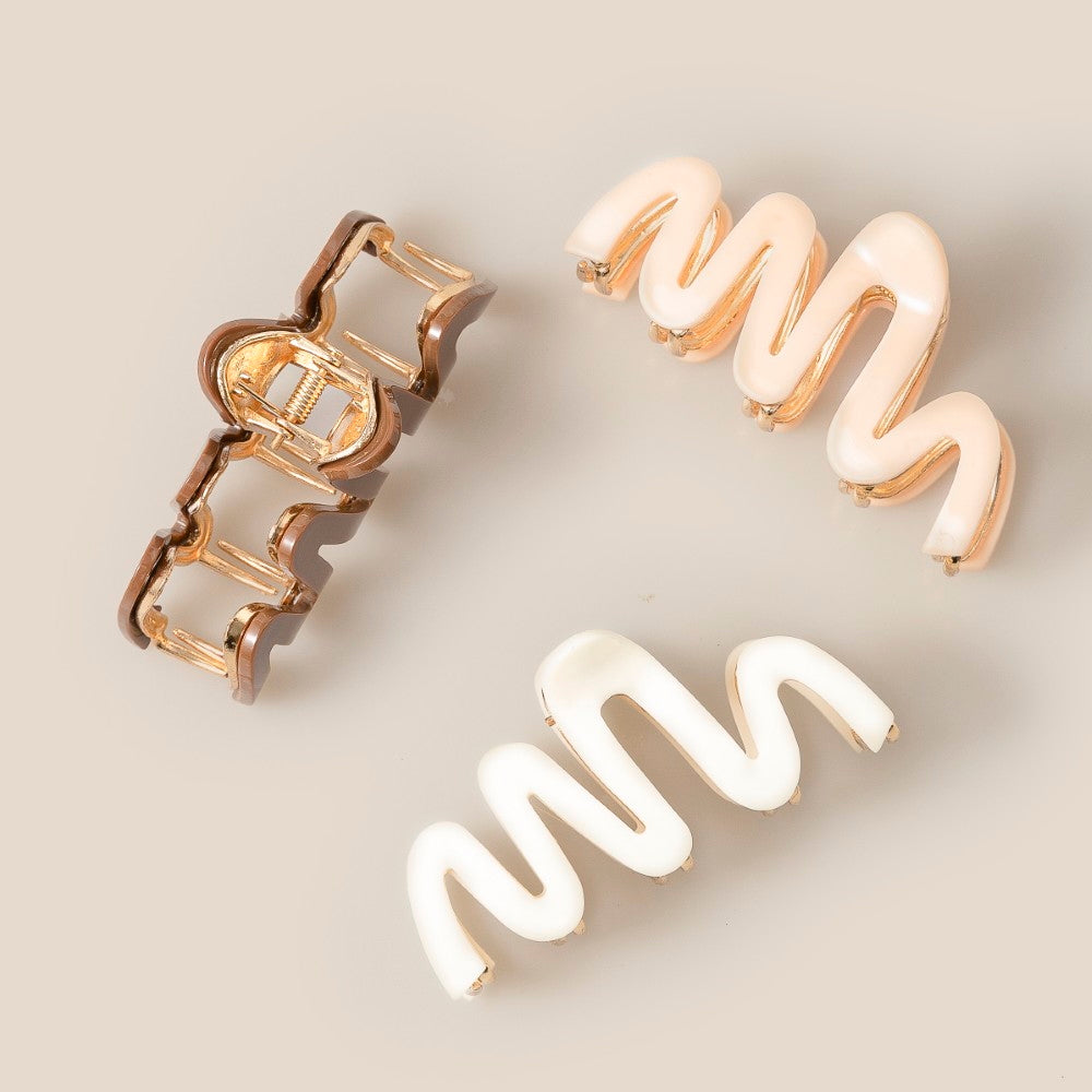 The South Moon Under Wavy Assorted Clips in Brown Multi feature three modern hair accessories—a rounded gold-tone clip and two wavy clips in pearly white and pale pink—arranged on a beige surface.